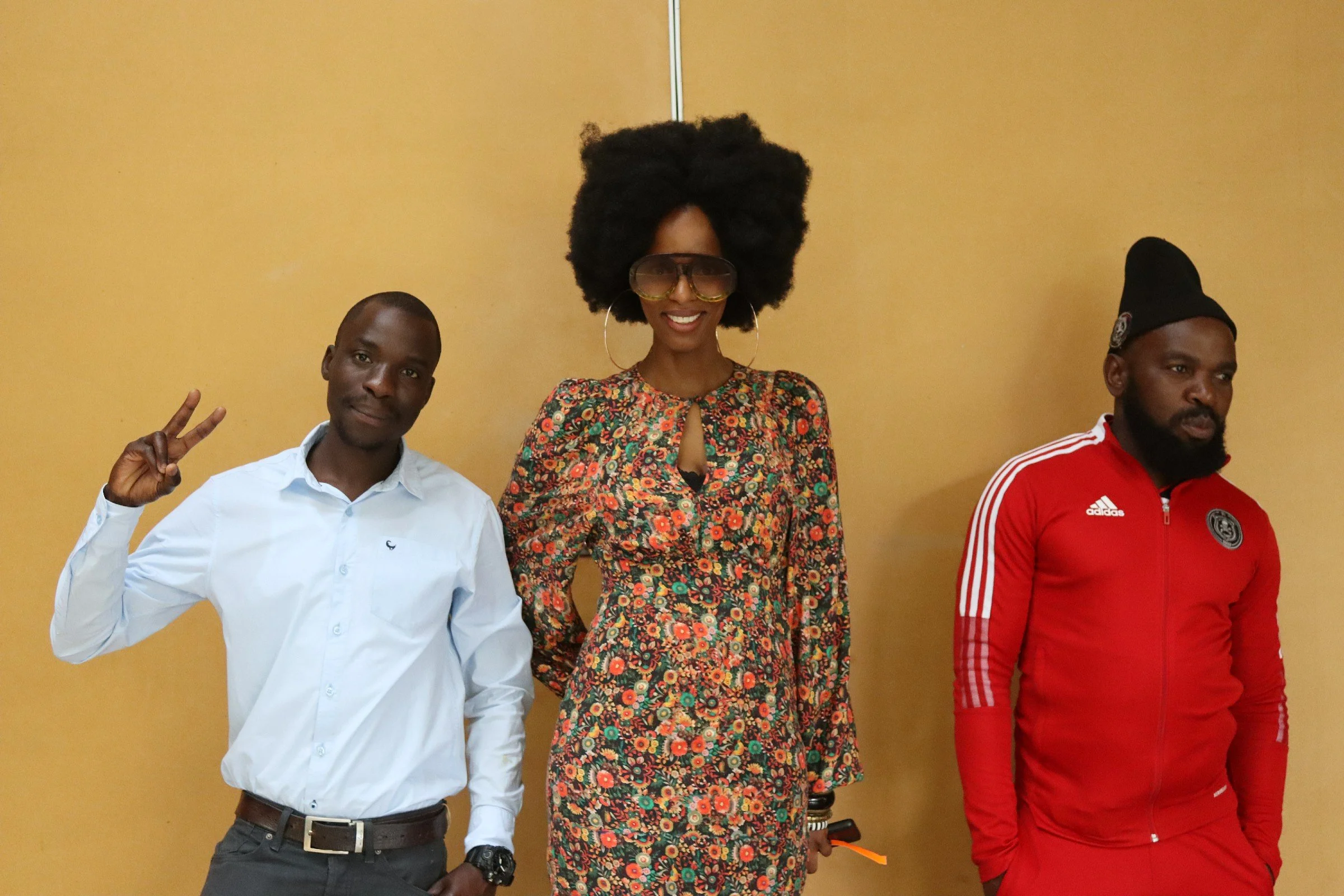 Three people standing against a beige wall, with the person on the left making a peace sign. The woman in the middle has a large afro hairstyle, wearing sunglasses and a floral dress. The man on the right is wearing a red sports jacket and a black be