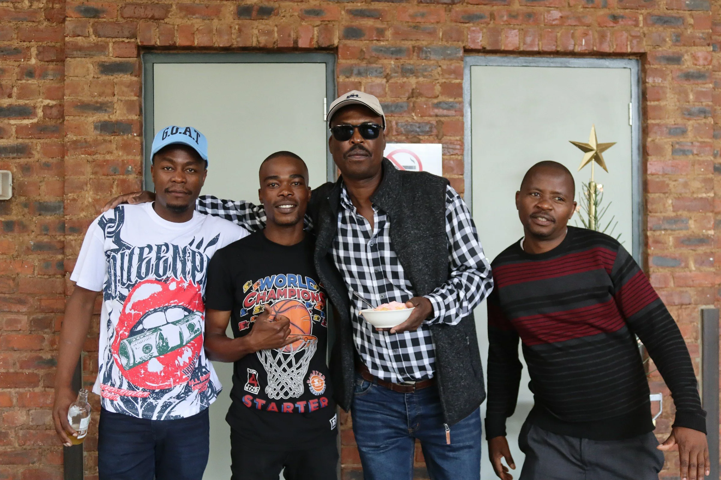 Five men standing in front of a brick wall, posing for a photo. One is holding a bowl of food, and another has a bottle in hand. All are wearing casual clothing, and one is wearing sunglasses and a cap.