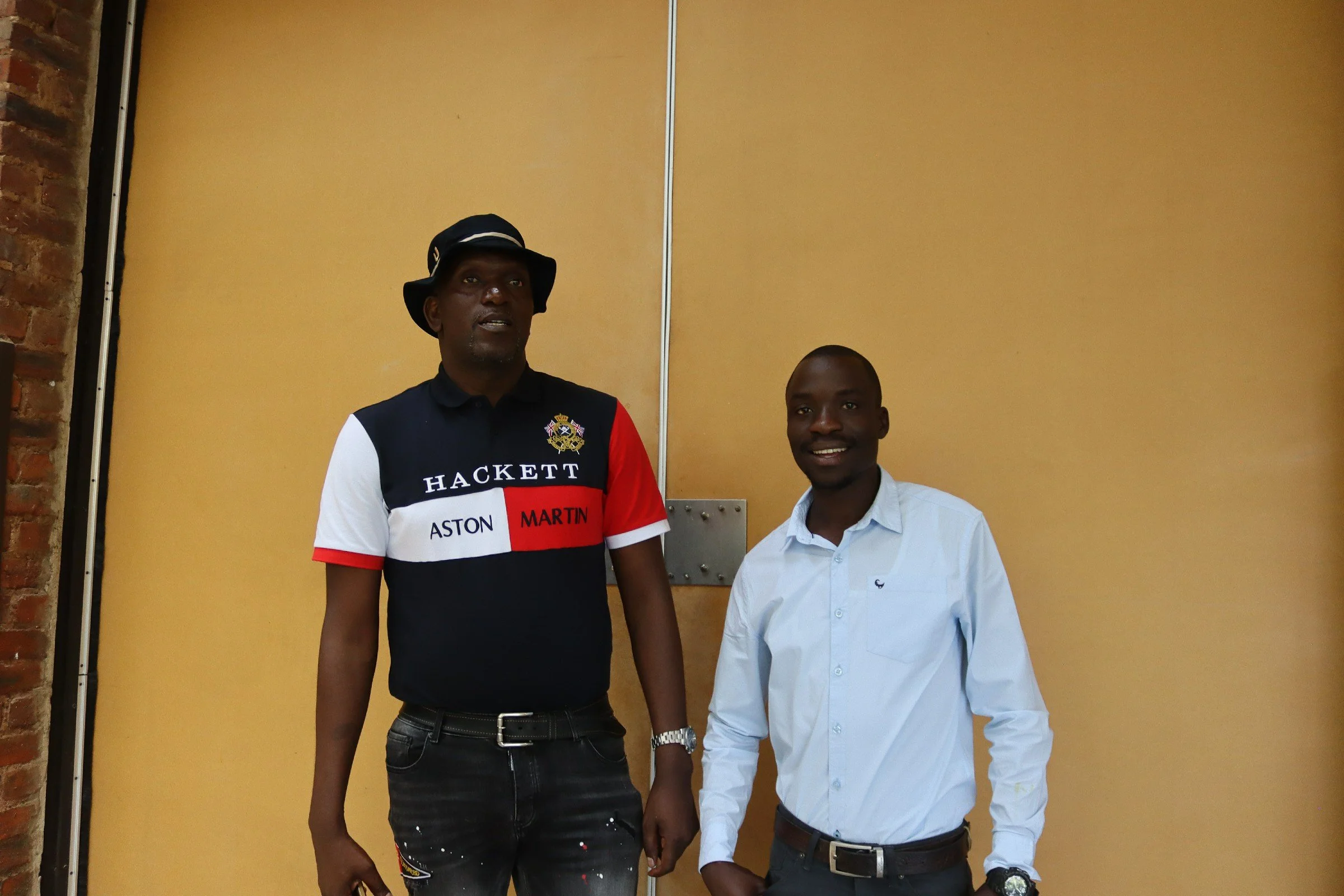 Two men standing indoors against a yellow wall, holding hands. One is wearing a black bucket hat and a colorful polo shirt with branding, the other is wearing a light blue dress shirt and dark pants.