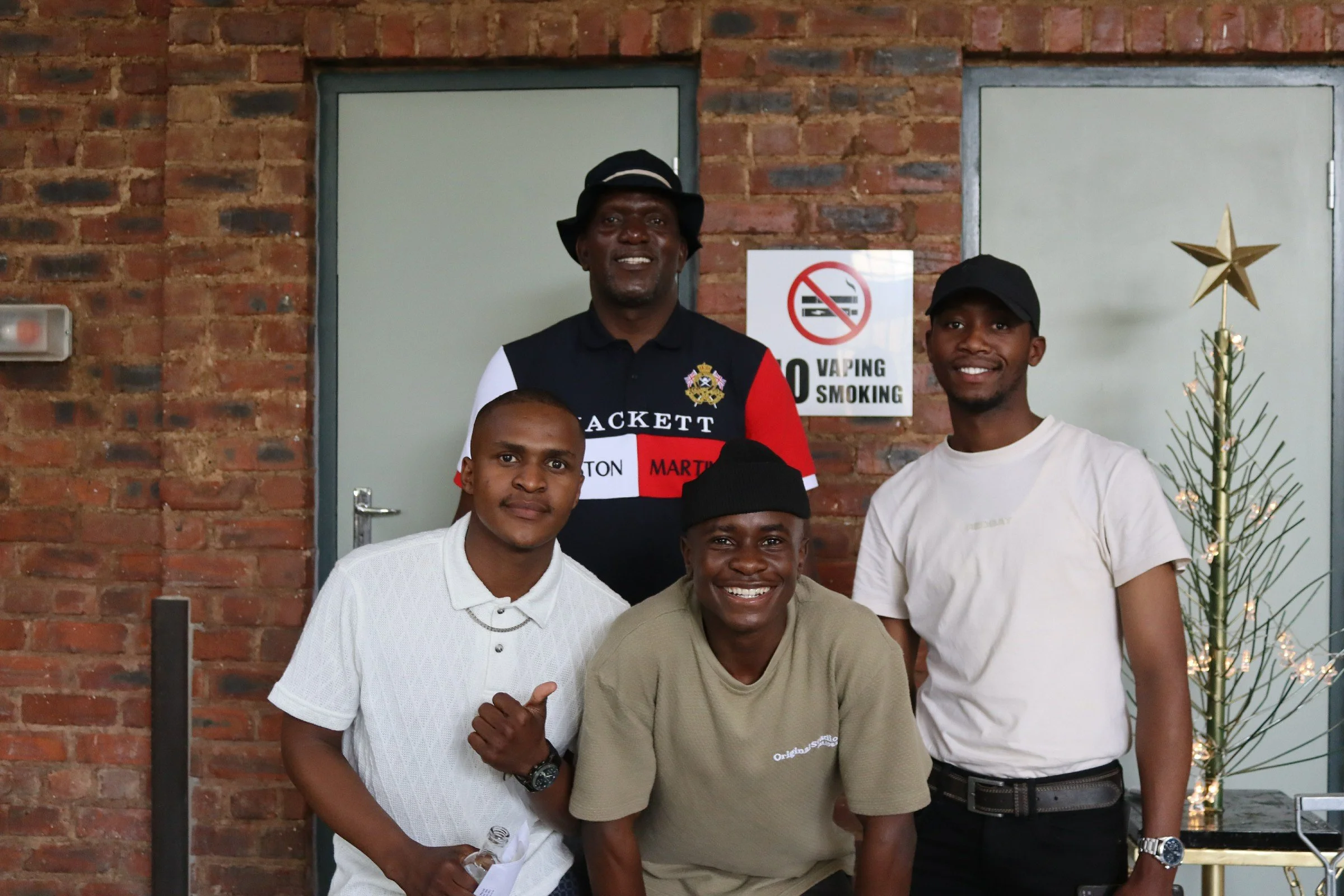A group of five young men smiling and posing for a photo indoors. They are standing in front of a brick wall with a no smoking sign and a small decorated Christmas tree. The man on the left is wearing a white shirt, the man in the middle is wearing a