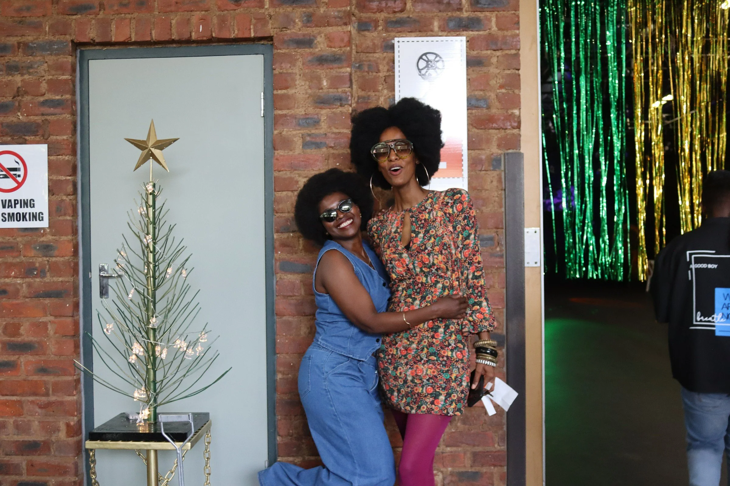 Two women with large curly hairstyles posing together indoors in front of a brick wall, with a small decorated Christmas tree nearby and gold and green hanging decorations to the side.