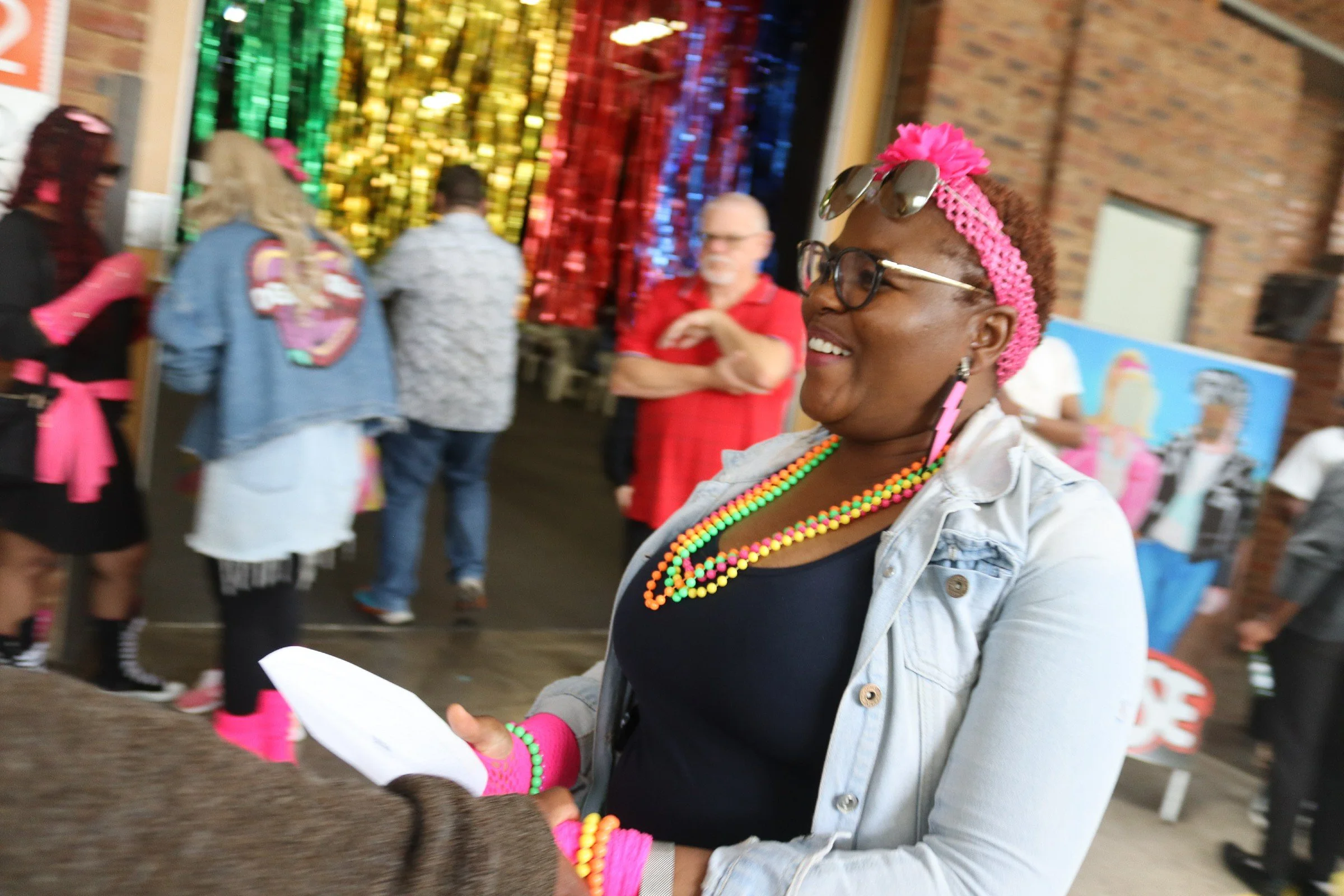 A woman wearing sunglasses, a pink headband with a pink flower, and colorful bead necklaces is smiling and talking to someone at a lively indoor event with people and Christmas decorations in the background.