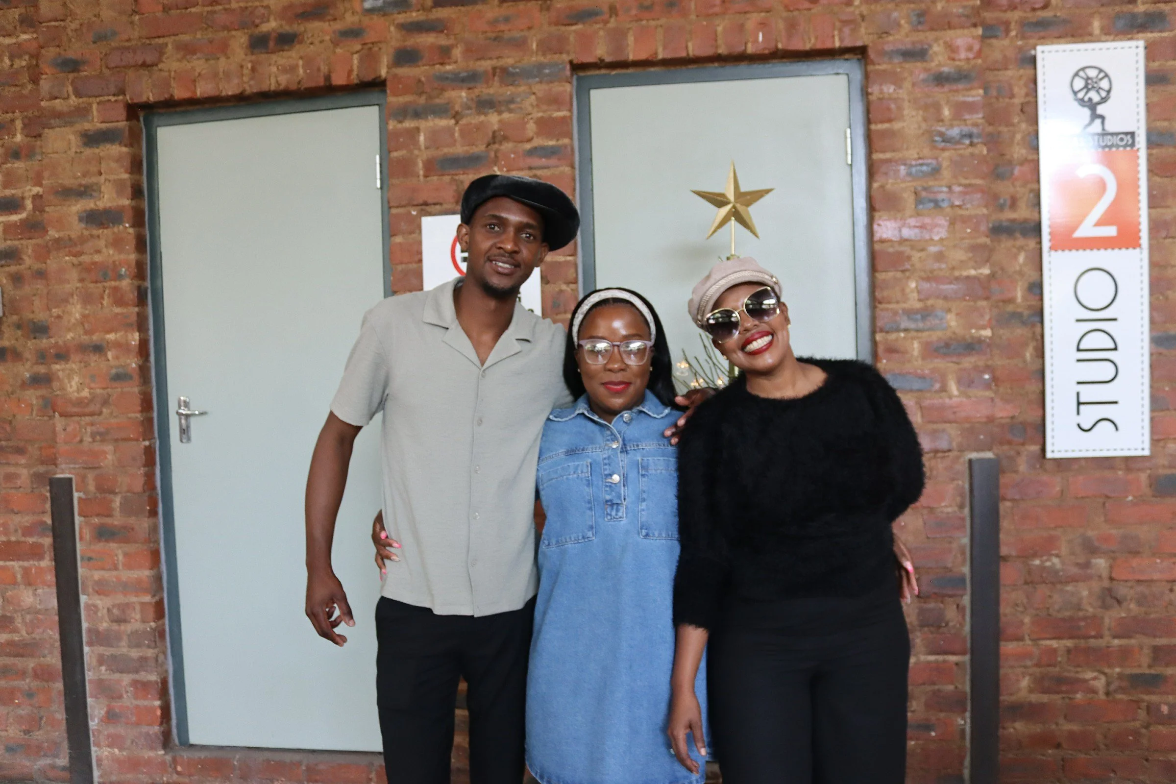 Three people standing together in front of a brick wall, smiling. The person on the left is a man wearing a beige shirt and a black beret. The middle person is a woman with glasses, wearing a denim jacket and a headband. The person on the right is a 