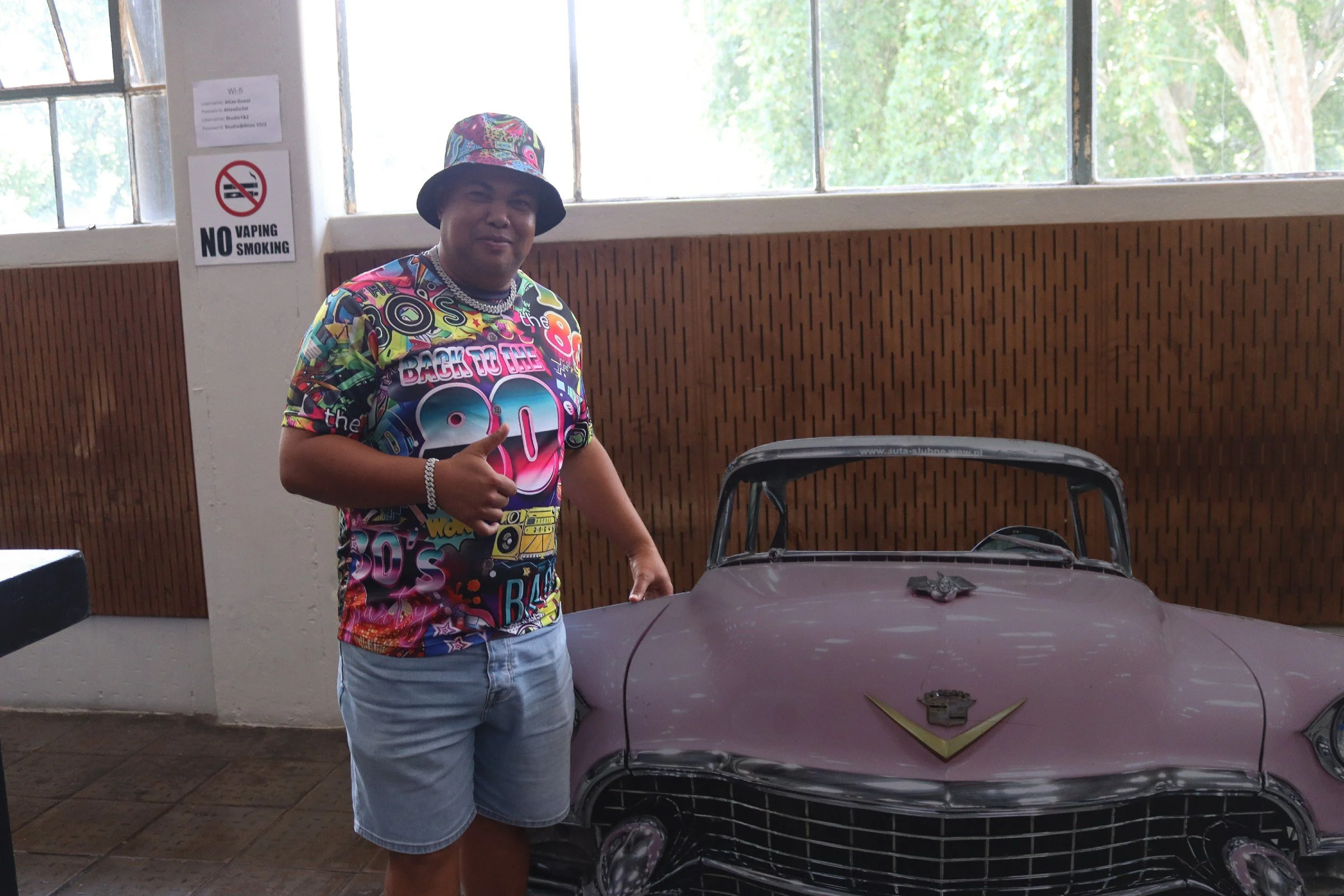A person wearing a colorful patterned bucket hat and a vibrant shirt with 'Back to the 80s' printed on it, giving a thumbs-up, standing next to a miniature car with Batman and Wonder Woman logos on the hood.