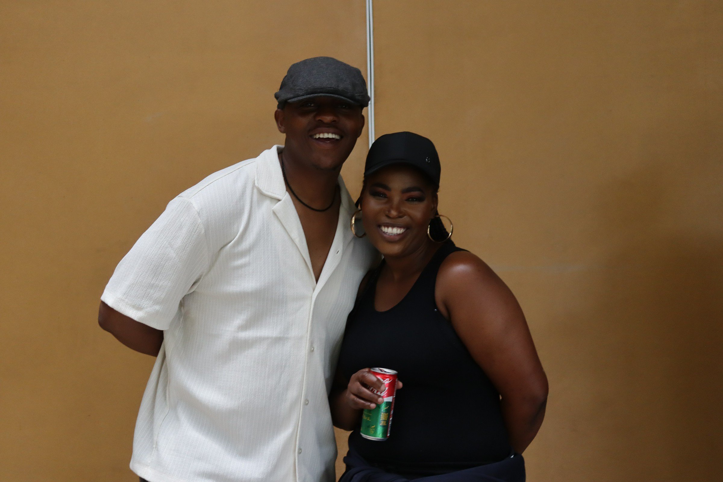 A man and a woman are standing together, smiling for the camera, against a plain brown wall. The man is wearing a gray flat cap and a white shirt, and the woman is wearing a black cap, gold hoop earrings, and a black sleeveless top, holding a red and
