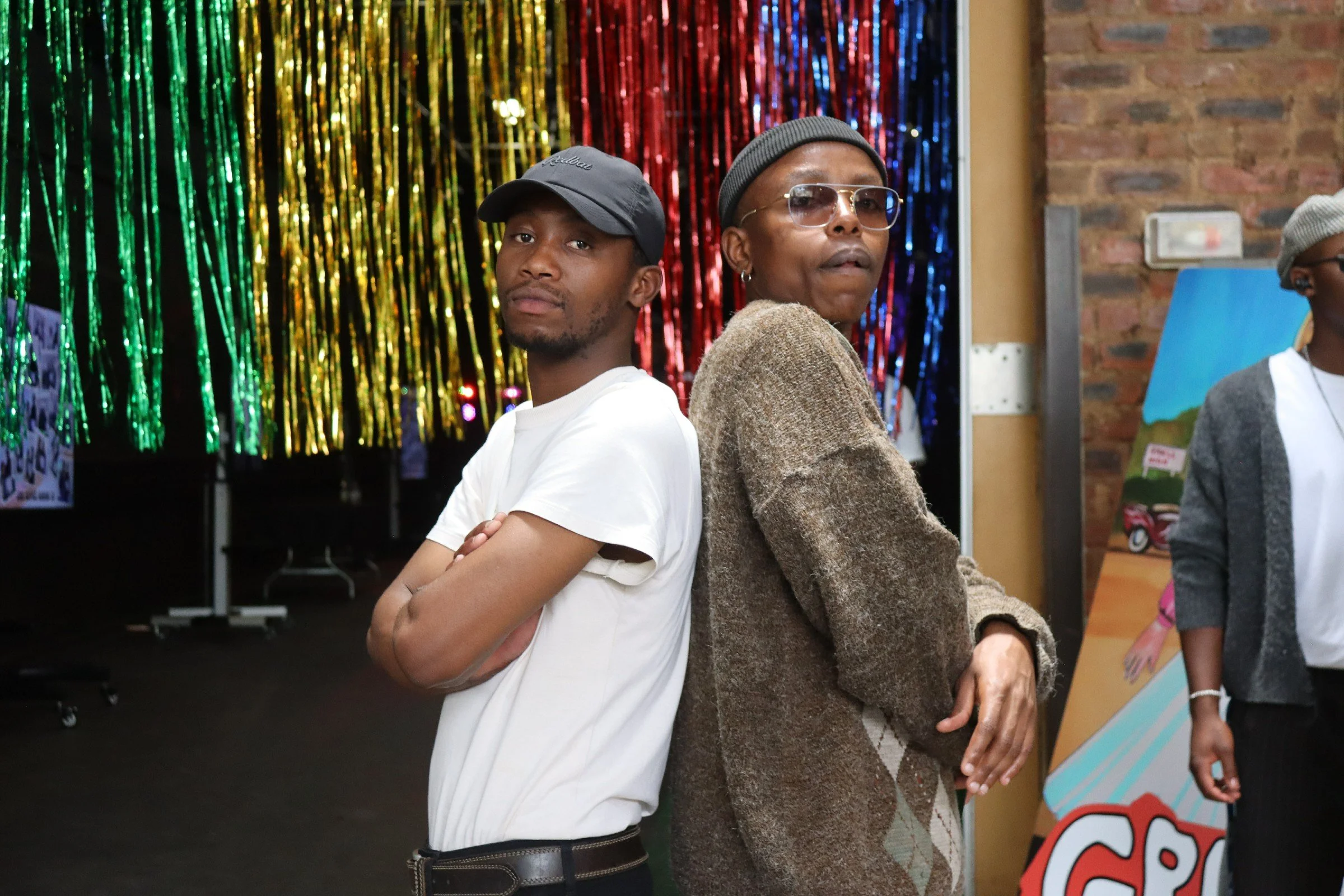 Two men with arms crossed stand back to back in a festive indoor setting with colorful streamer curtains in the background. One is a young man with a baseball cap and white t-shirt, the other is an older man with glasses, earrings, a beanie, and a br