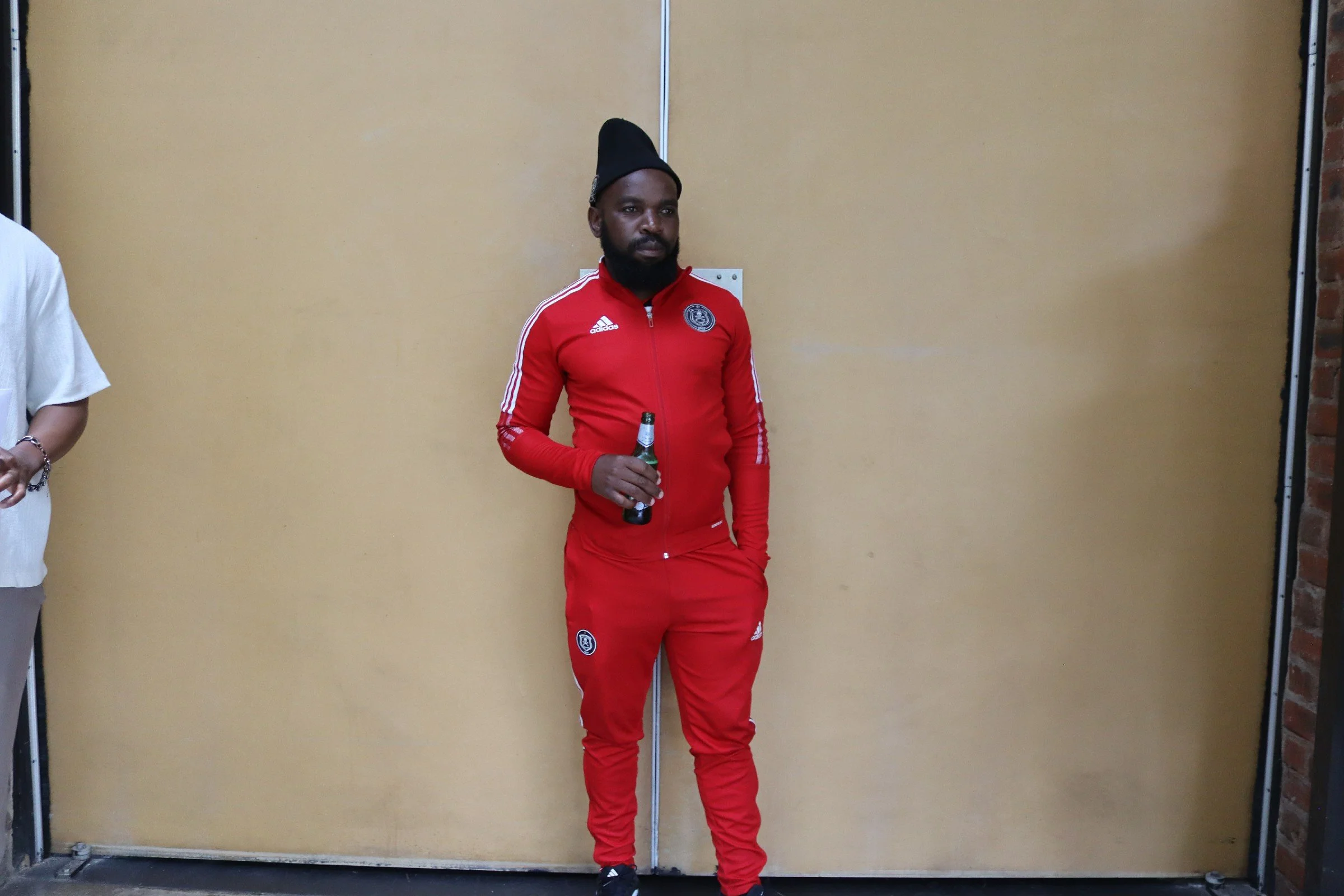 A man with a dark beard wearing a red Adidas tracksuit and a black hat, holding a bottle, standing against a beige wall.
