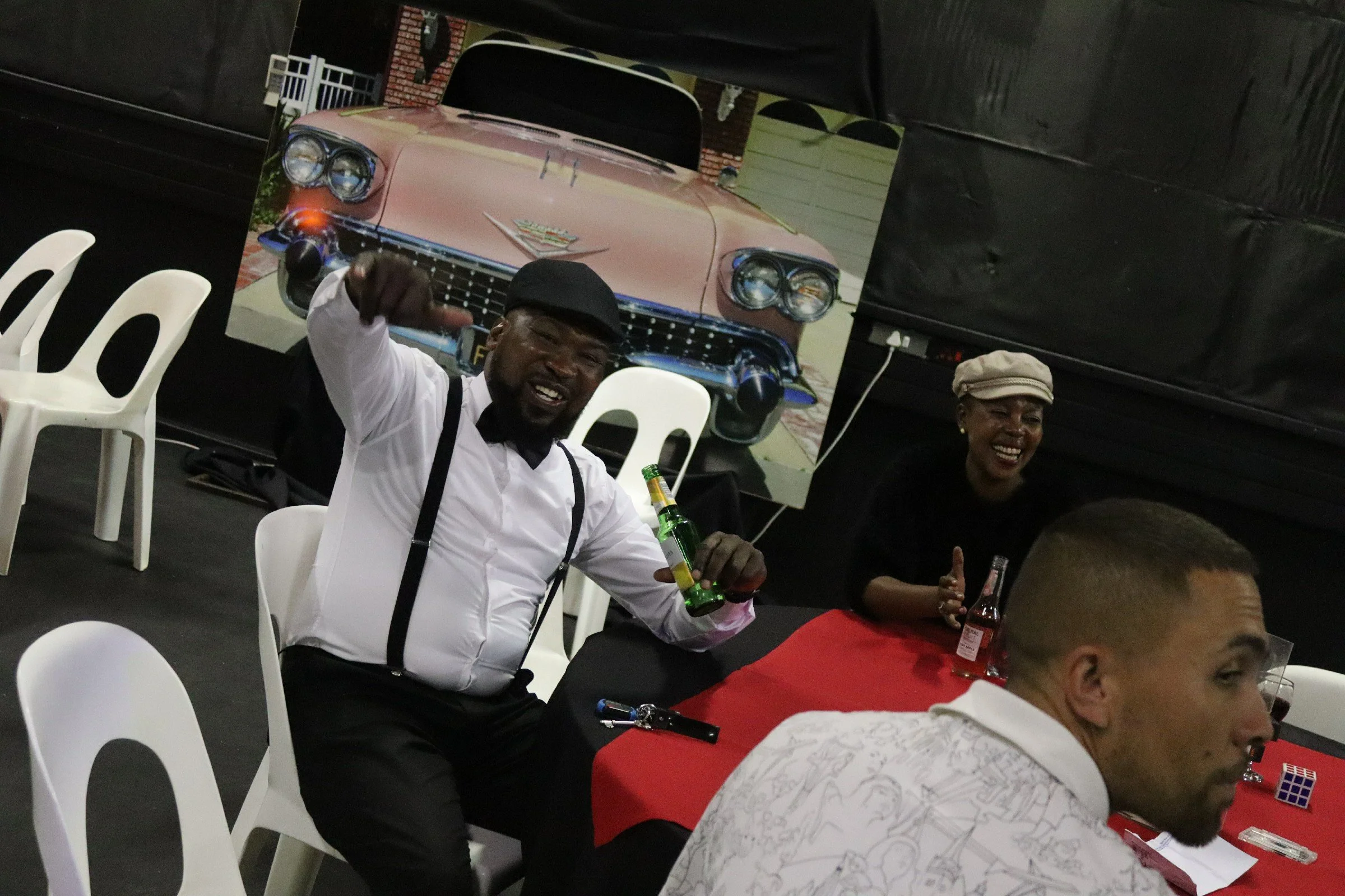 People sitting at tables with white and red tablecloths, some holding bottles and giving thumbs up, in an indoor setting with a large poster of a vintage pink car in the background.