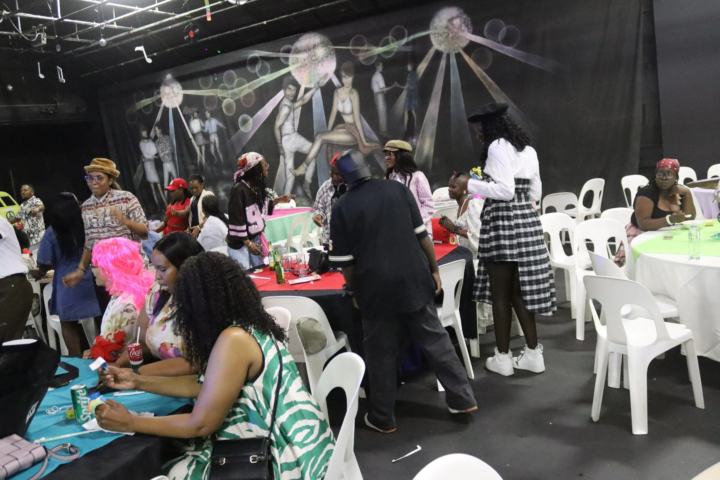 People gather at a celebration or party in an indoor venue with a dark wall featuring a mural of figures and abstract designs. Some guests are seated at tables with drinks, while others are standing, chatting, or walking around. The atmosphere appear