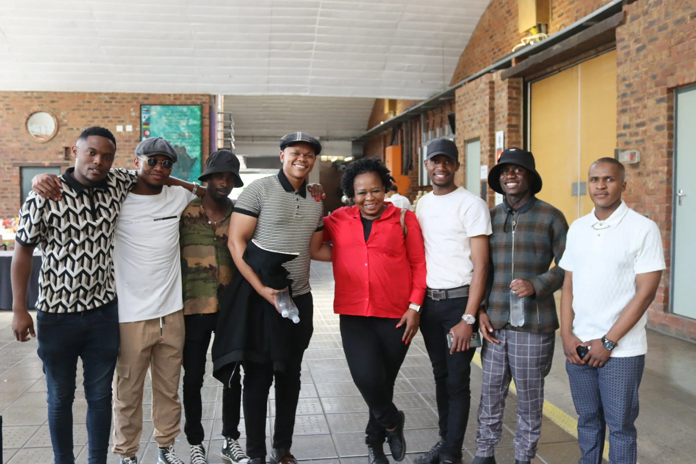 Group of nine diverse people standing together in an indoor setting, smiling at the camera, with a brick wall and digital screen in the background.