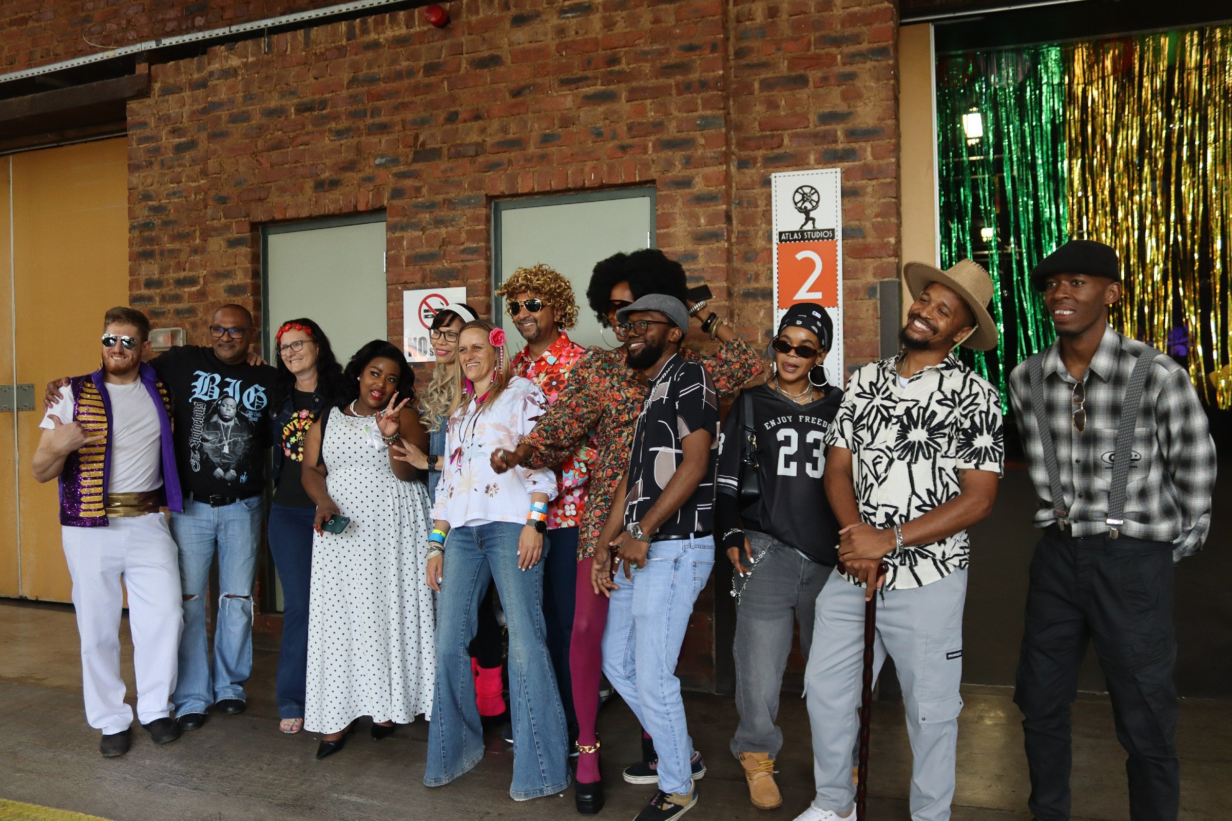 Group of diverse people smiling and posing together outside an industrial brick building, some wearing sunglasses and colorful clothing.