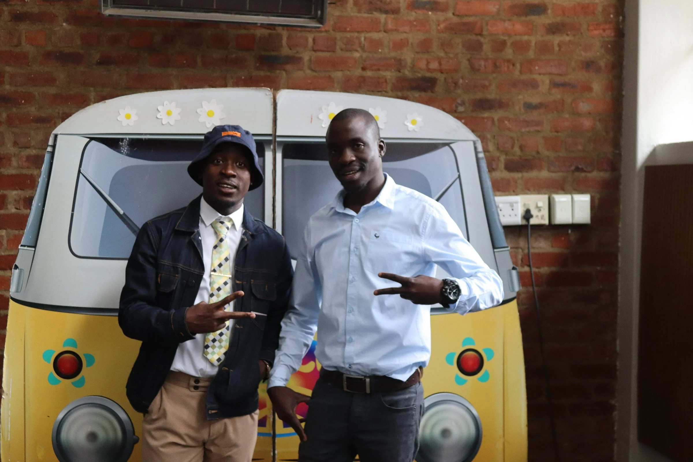 Two young men standing in front of a colorful speaker setup. The man on the left wears a dark jacket, beige pants, a patterned tie, and a bucket hat. The man on the right wears a light blue button-up shirt and dark pants, making a peace sign with his