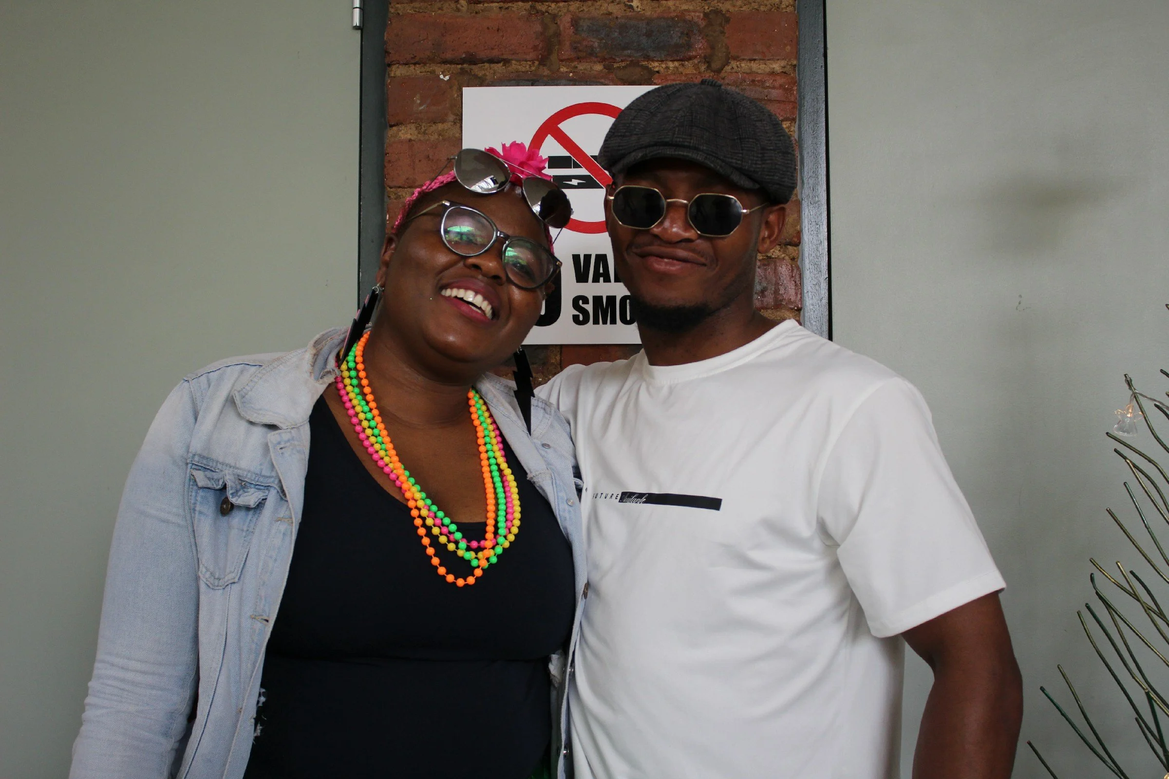Two people smiling, posing together indoors in front of a brick wall with a 'No Smoking' sign, both wearing sunglasses and casual clothing.