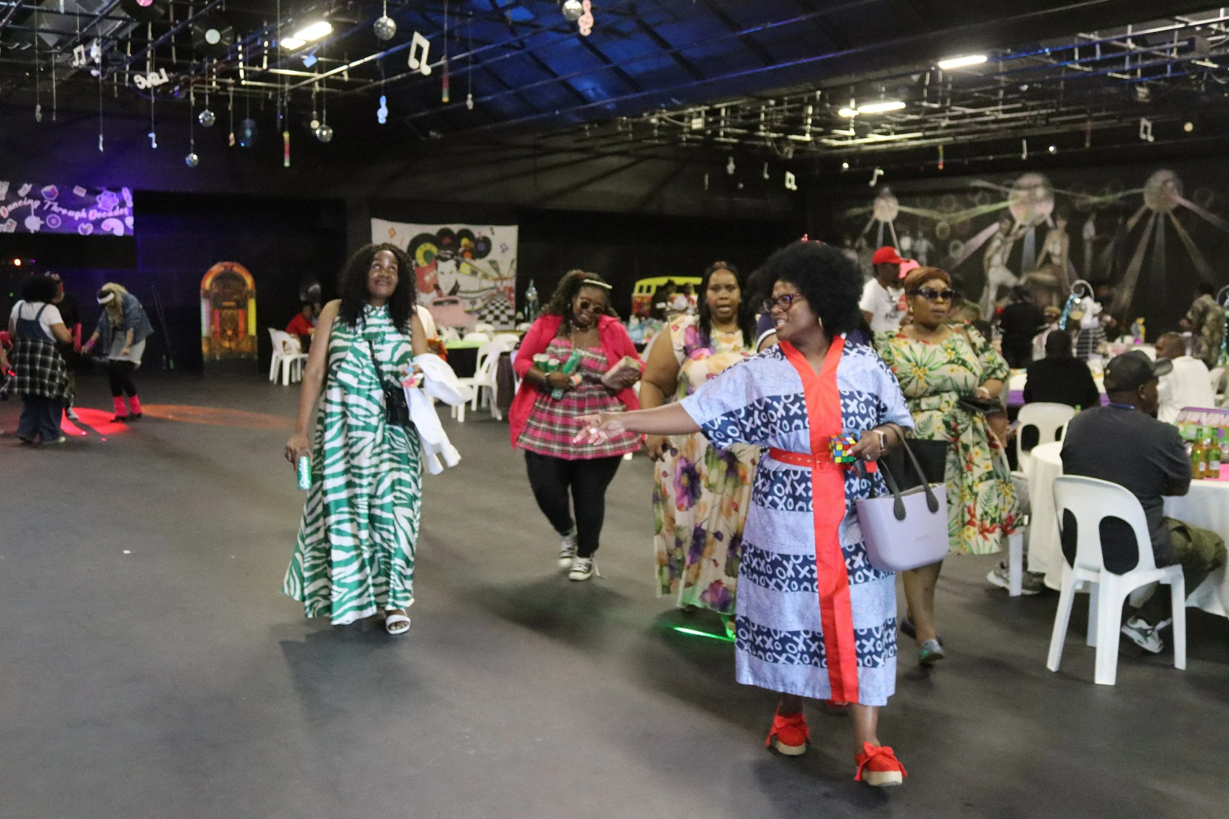 Group of women in colorful dresses dancing and socializing in a decorated indoor space with tables, chairs, and wall art.