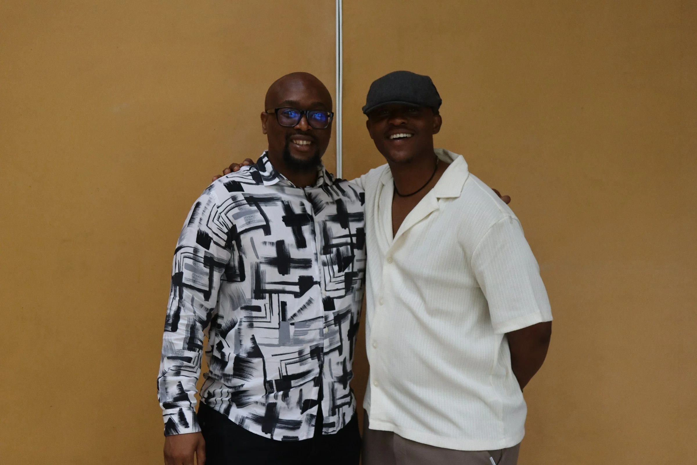 Two men smiling and standing close together in front of a plain beige wall, with one man wearing glasses and a patterned shirt, and the other wearing a cap and a light-colored shirt.