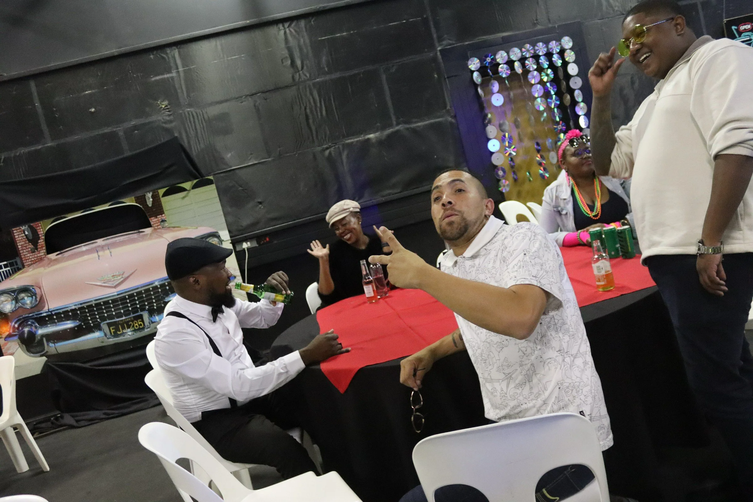 People socializing at a gathering with vintage cars in the background. One person is wearing a white shirt, bowtie, and black hat, sipping from a green bottle. Another man with tattoos, wearing glasses and a white patterned shirt, is sitting at a tab