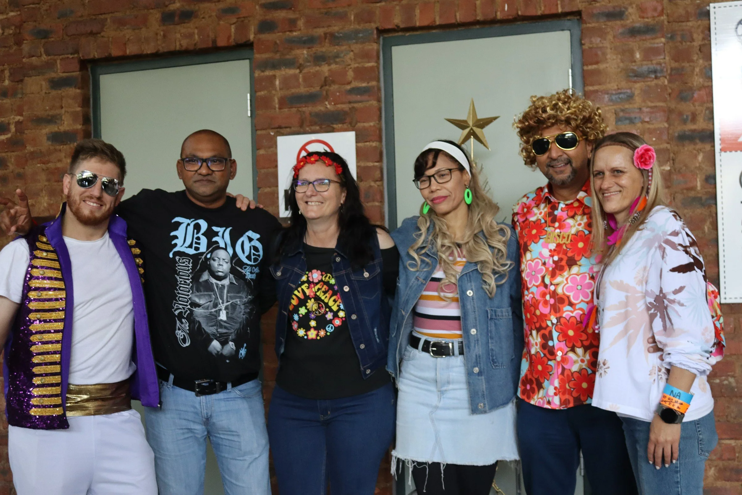 Group of six people dressed in colorful, retro, or hippie-style clothing posing together against a brick wall with two large gray doors, some wearing sunglasses, and smiling at the camera.