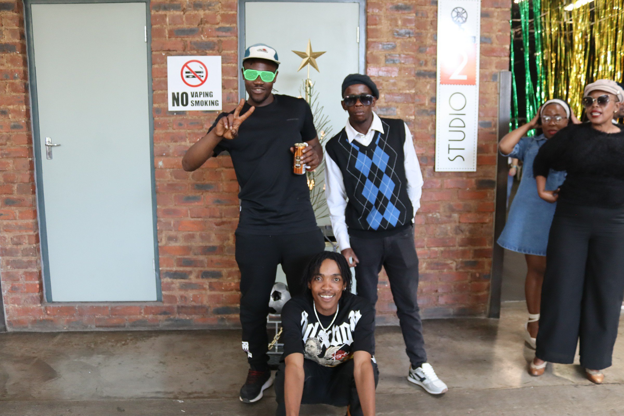 A group of young people posing in front of a brick wall with a sign that says "No Vaping Smoking" and a Christmas tree in the background. They are dressed casually, with some wearing sunglasses and hats, and smiling or making peace signs.