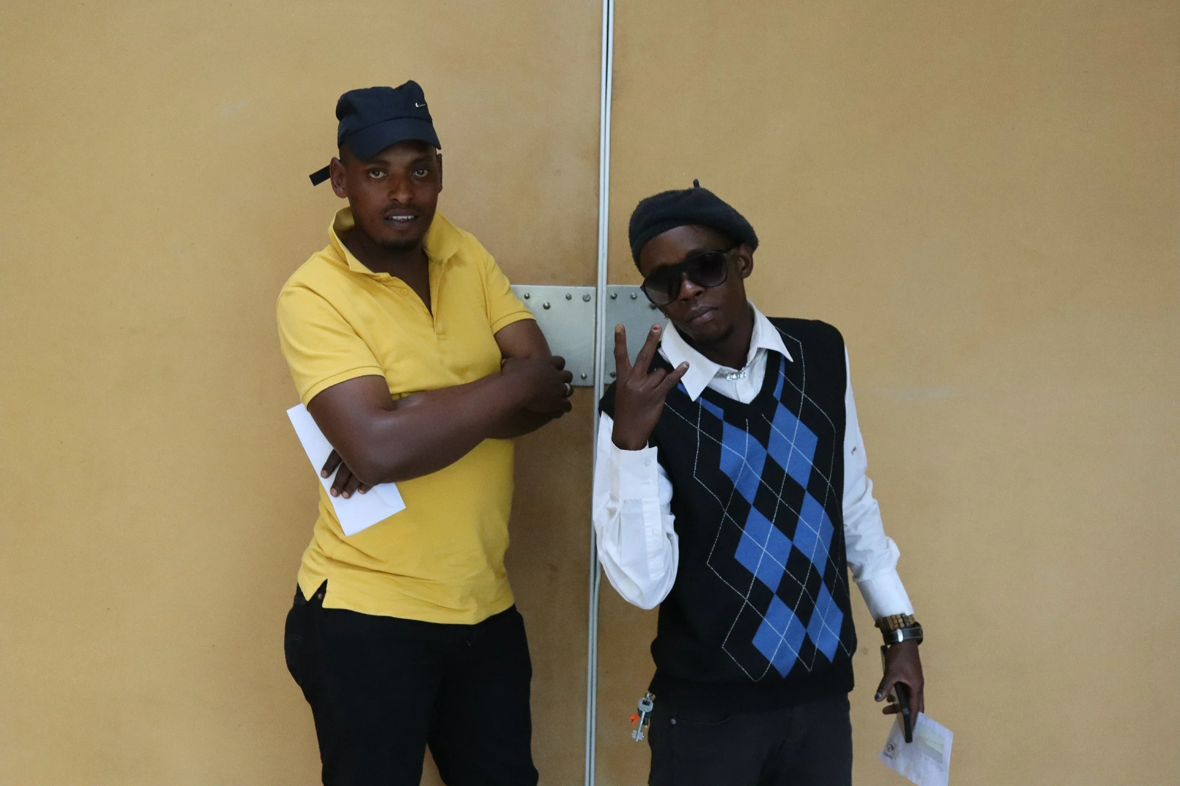 Two young men standing against a yellow wall, one dressed in a yellow shirt and black pants with a dark cap, the other in a white shirt with a black and blue argyle vest, wearing sunglasses and a black beanie, making a peace sign with his fingers.