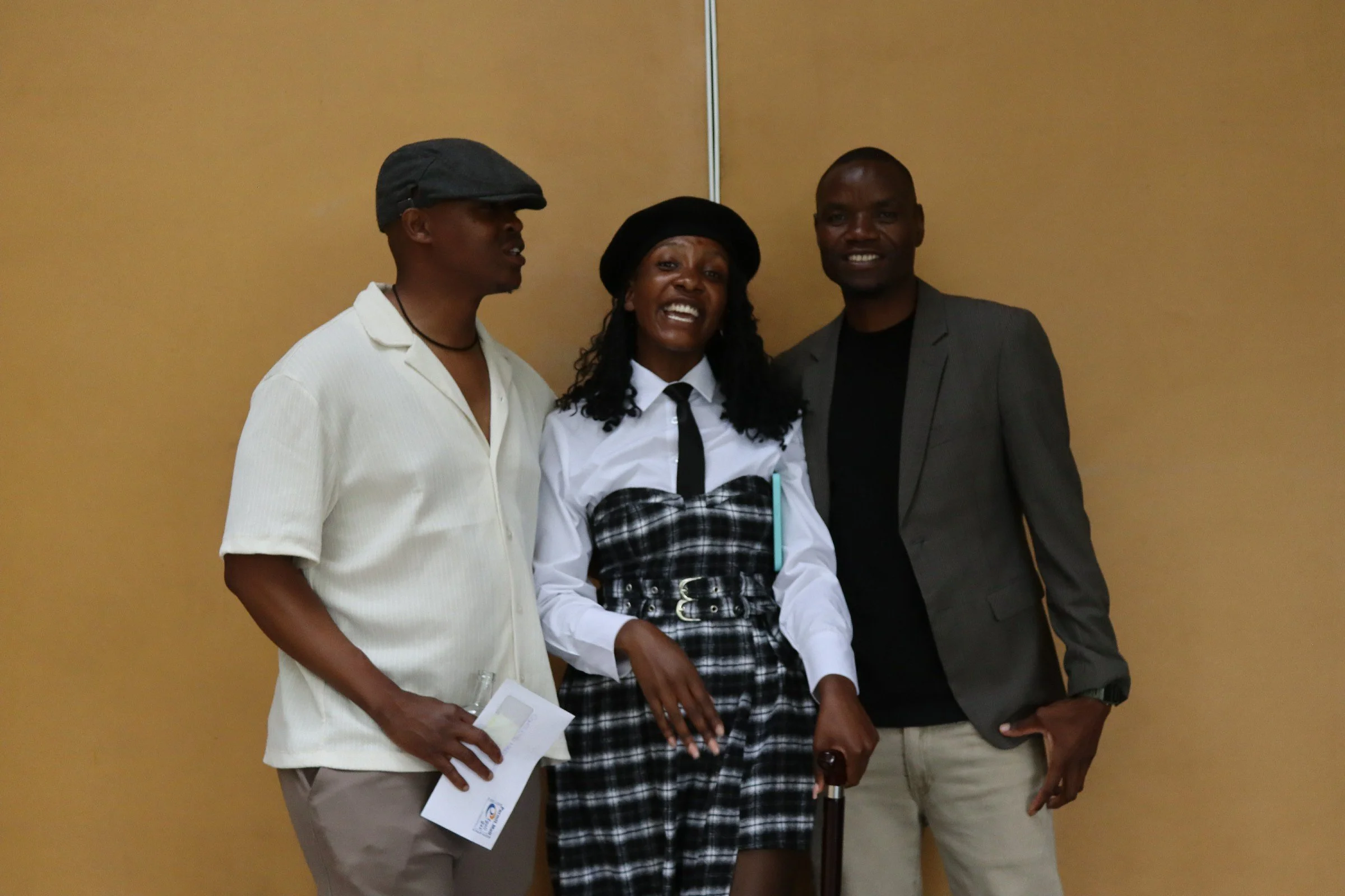 Three people standing against a beige wall, smiling and looking at the camera. The person on the left is wearing a white shirt and a gray cap, holding a paper. The person in the middle is a woman with curly hair, wearing a white shirt with a black ti