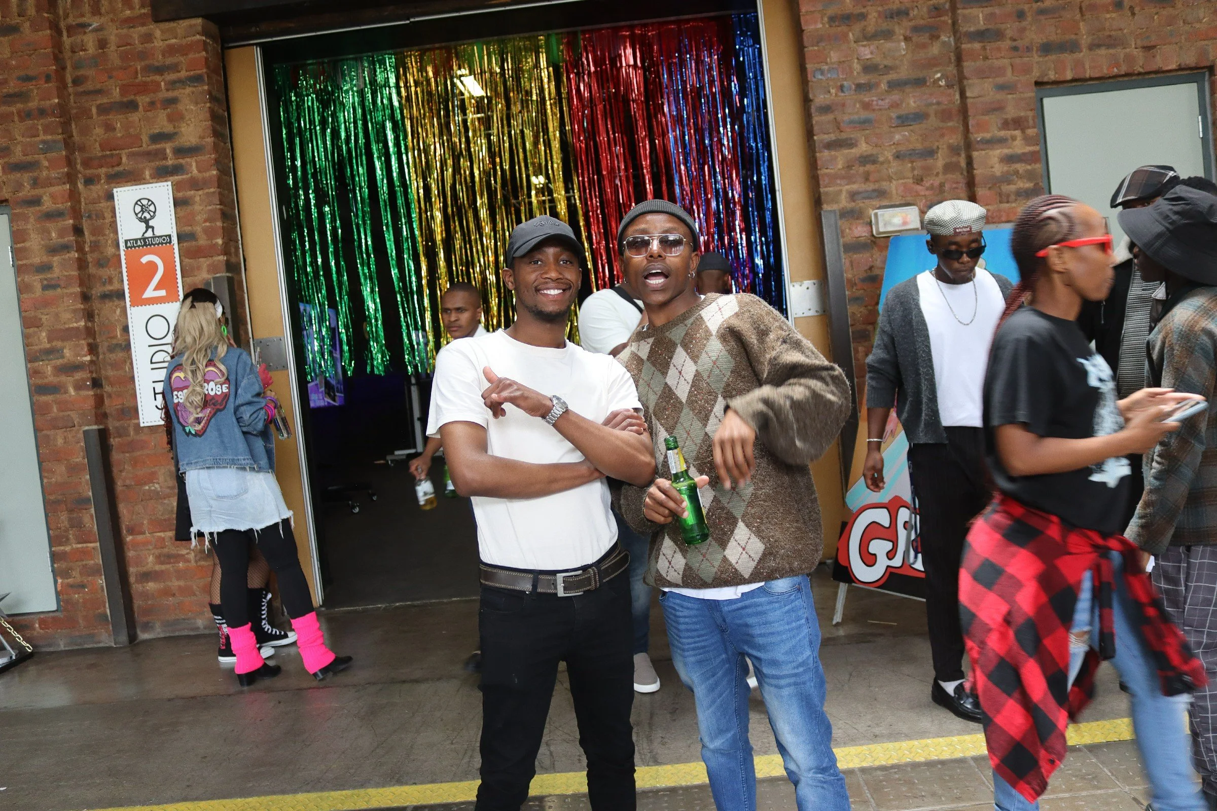 People gathered outside at an event, some smiling and chatting, with a colorful metallic curtain background behind them.