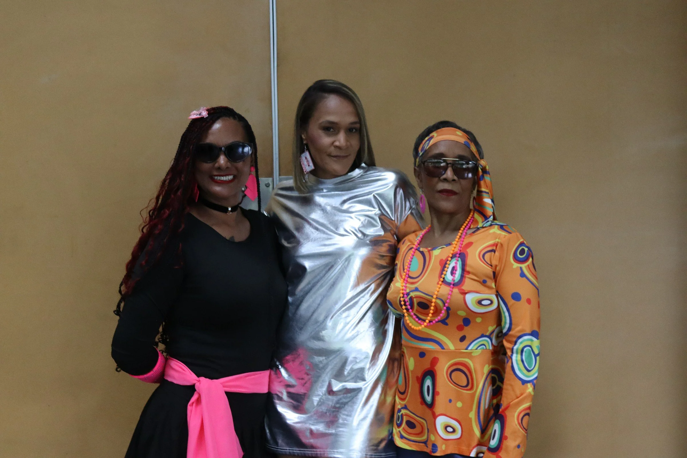 Three women standing together against a plain yellow wall. The first woman on the left has dark skin, long braided hair with red highlights, wearing large black sunglasses, a black top, pink gloves, and a pink belt. The middle woman has light skin, s