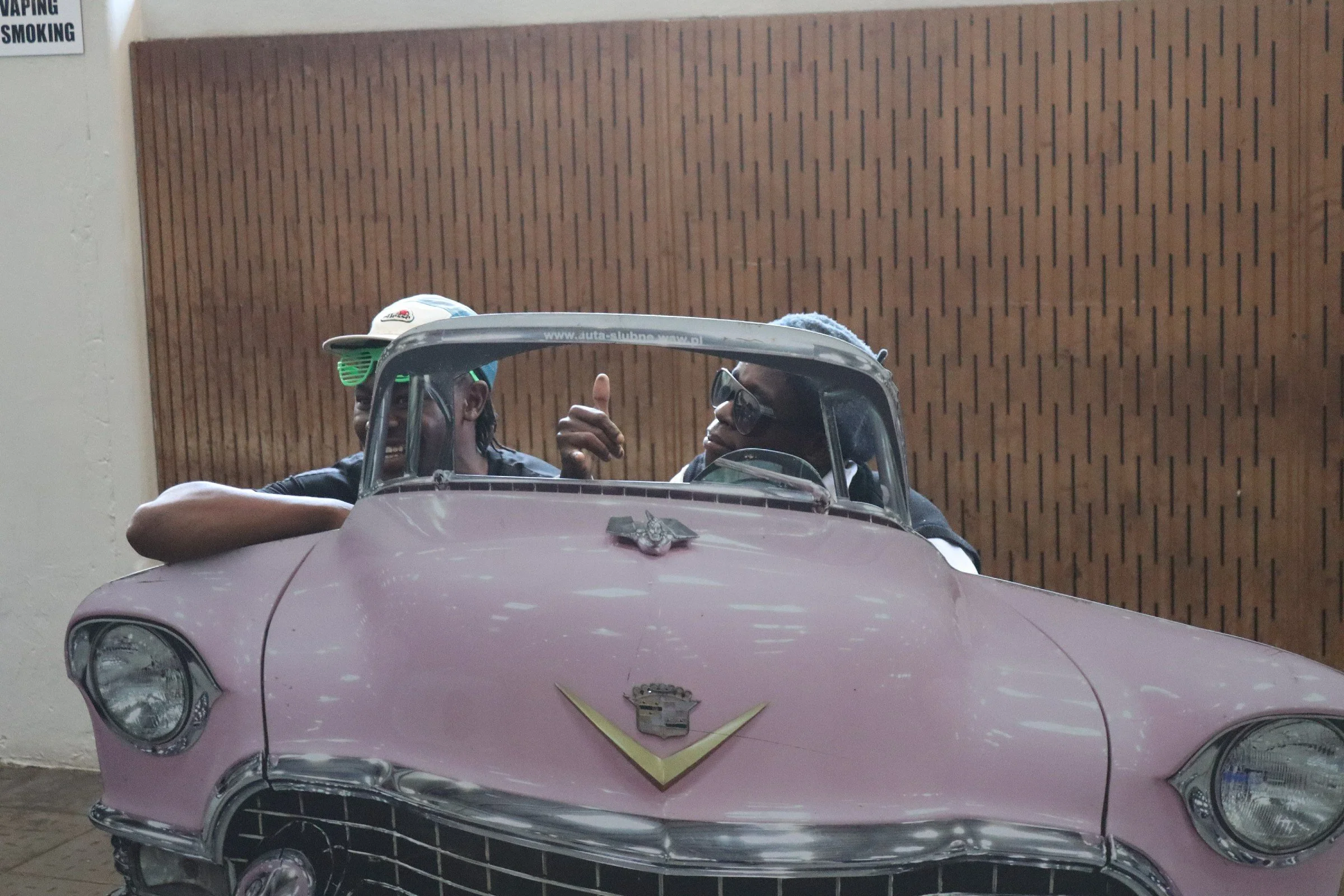 Two people sitting inside a pink Cadillac with a vintage Cadillac emblem on the front, with one giving a thumbs-up. They are wearing sunglasses and hats, in front of a wooden wall, with a sign that says 'No Vaping or Smoking' on the wall.