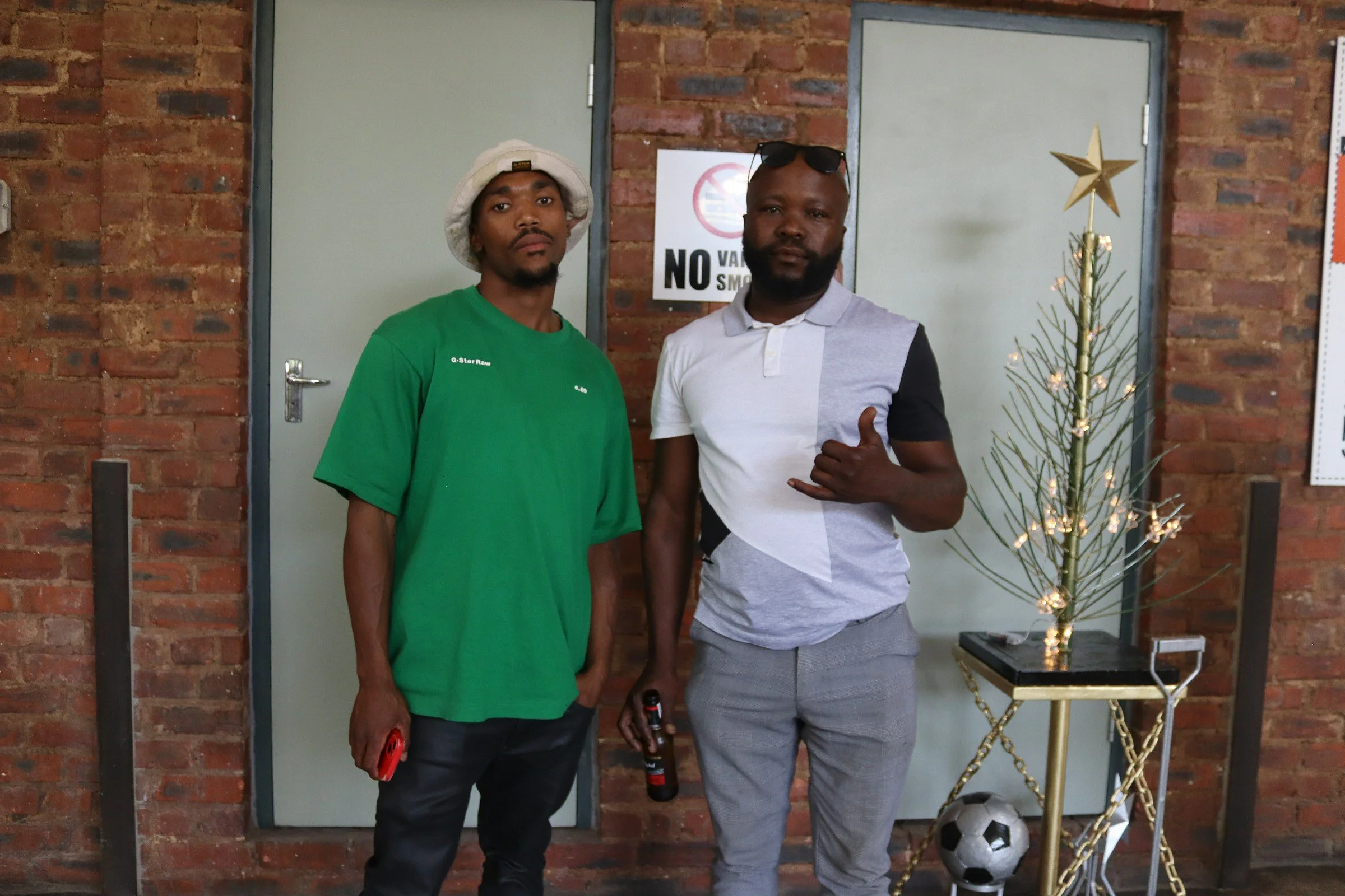 Two men standing indoors with a Christmas tree and a soccer ball. The man on the left wears a green t-shirt and a white bucket hat, holding a red object. The man on the right wears a gray and black polo shirt, sunglasses on his head, and is making a 
