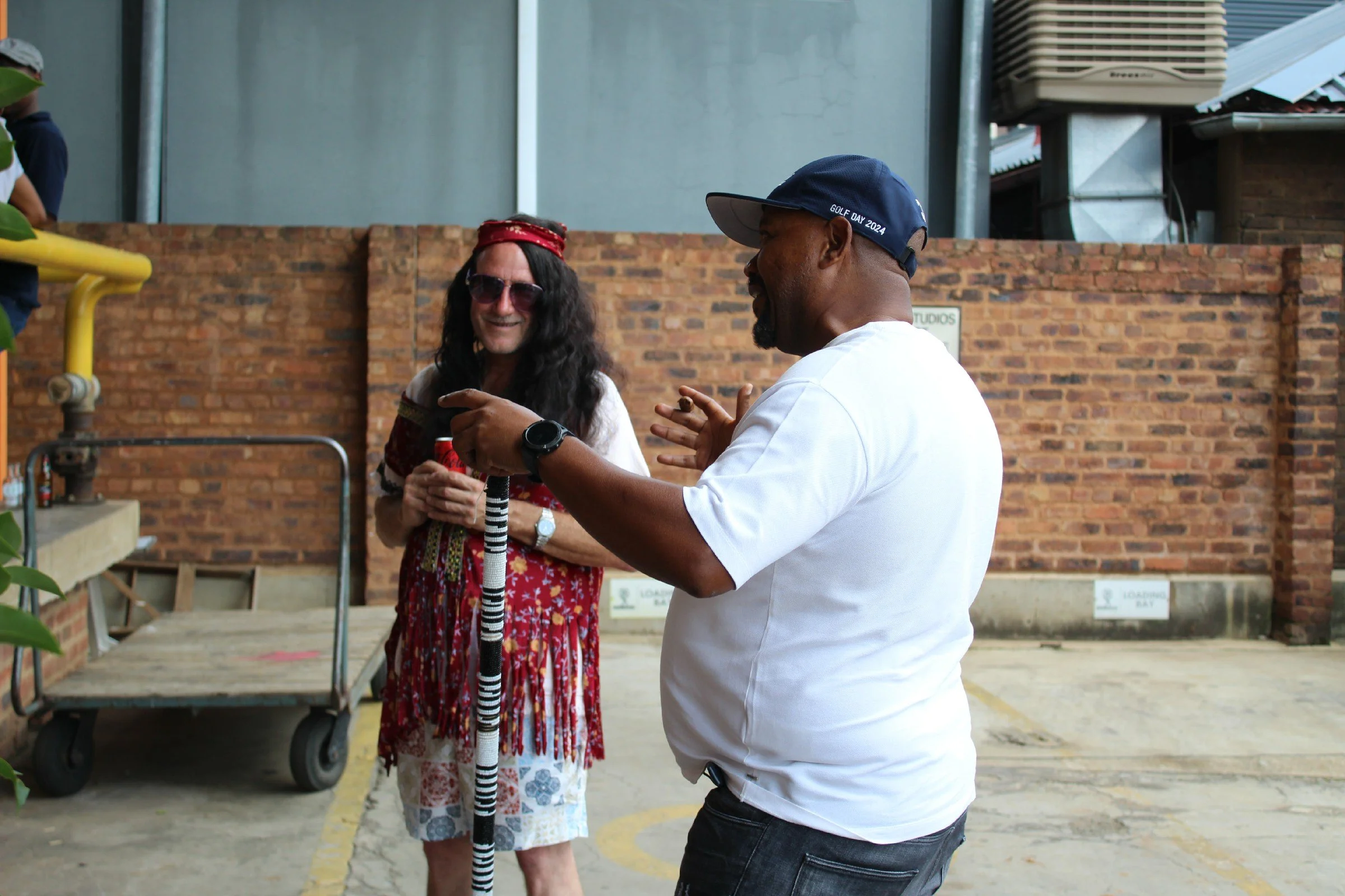 Two people are having a conversation outdoors against a brick wall. One person is dressed in a colorful outfit with a red headband and sunglasses, holding a striped staff. The other person is wearing a white shirt and a navy blue cap that reads 'GOLF