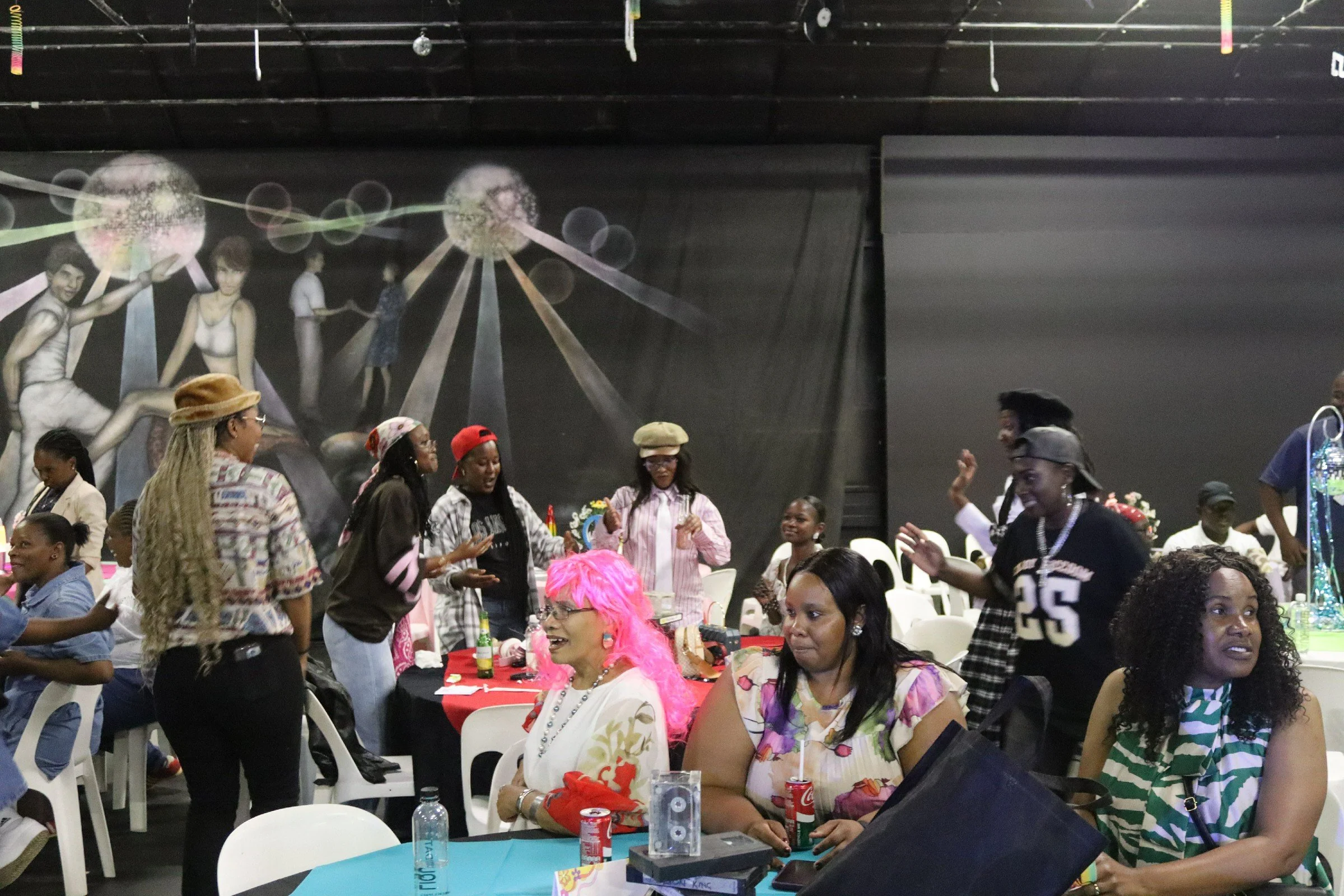 People gathered at a party or celebration, some standing and some seated at tables, with a large mural or painting of women and dandelion-like flowers in the background. Decorations and colorful attire indicate a festive atmosphere.