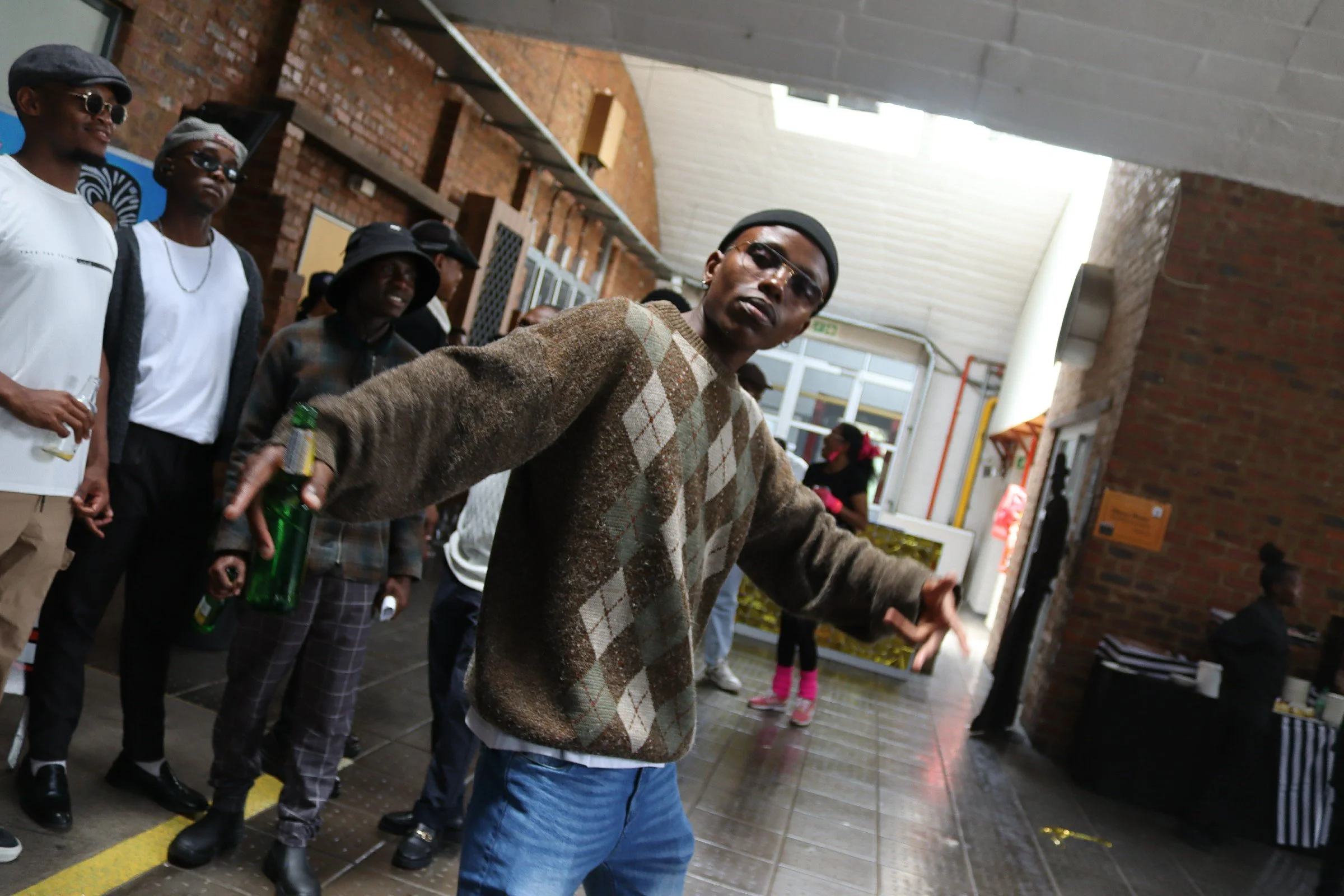 A group of young people standing in a line in an indoor space with brick walls, some wearing sunglasses and hats, with one person in front holding a green bottle and dressed in a brown plaid sweater and blue jeans.