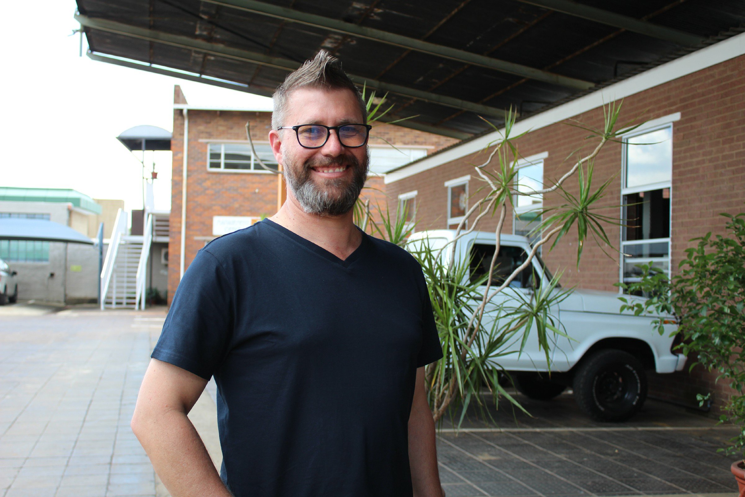 A smiling man with glasses and a beard standing outdoors in front of a building with brick walls and several windows, next to a potted plant, under a covered patio.