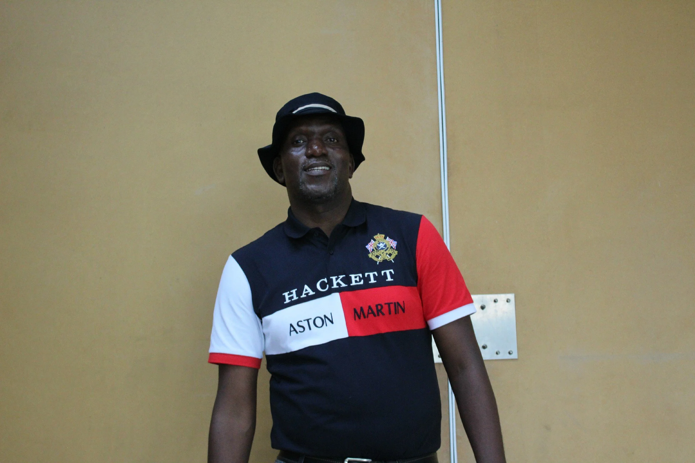 A man wearing a black bucket hat, a colorful short-sleeved shirt with the words 'HACKETT' and 'ASTON MARTIN' on it, standing in front of a beige wall with a metallic strip and electrical outlets visible.
