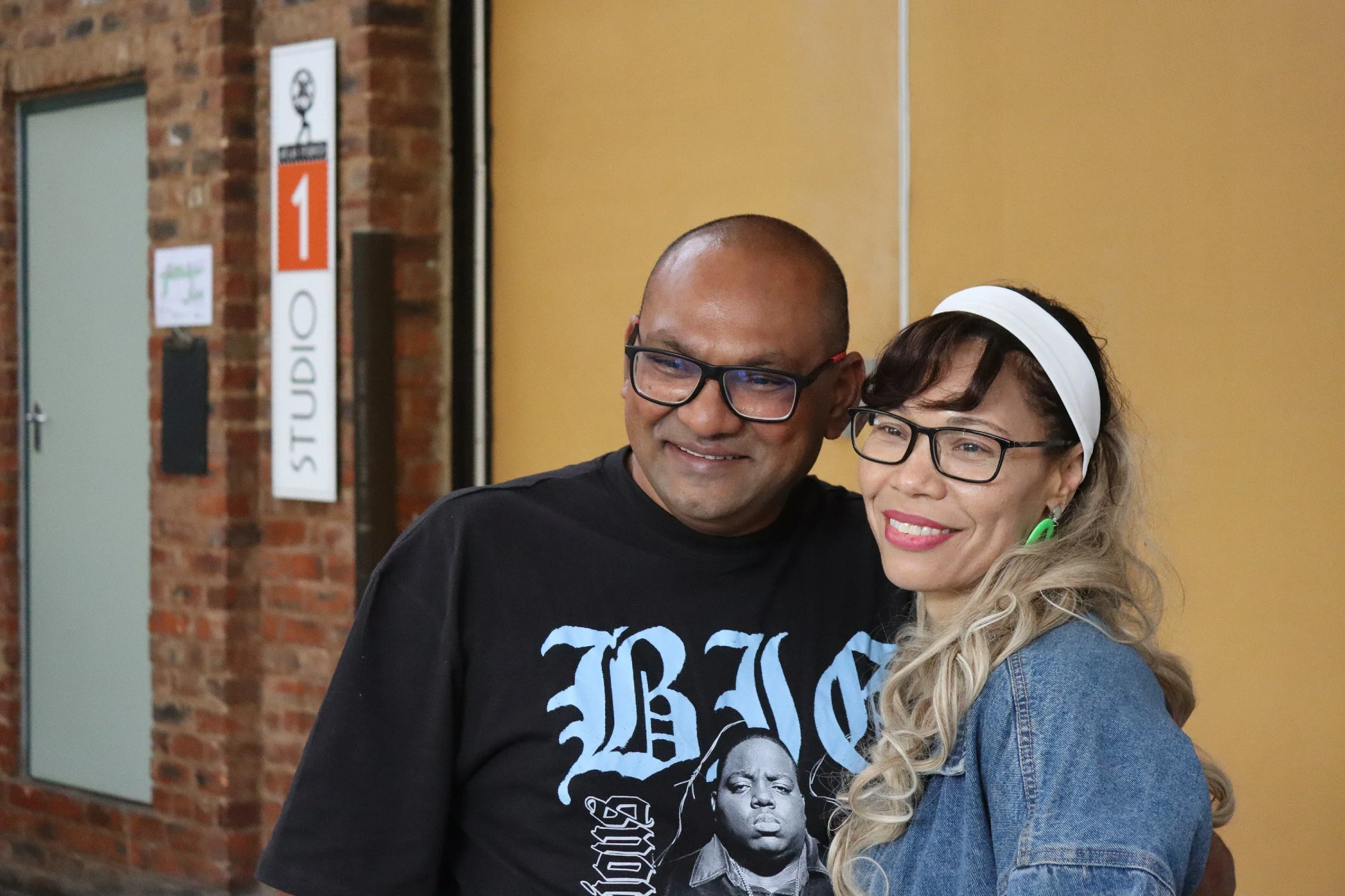 A man and a woman pose together, smiling, in an indoor setting with brick walls and a yellow partition in the background.