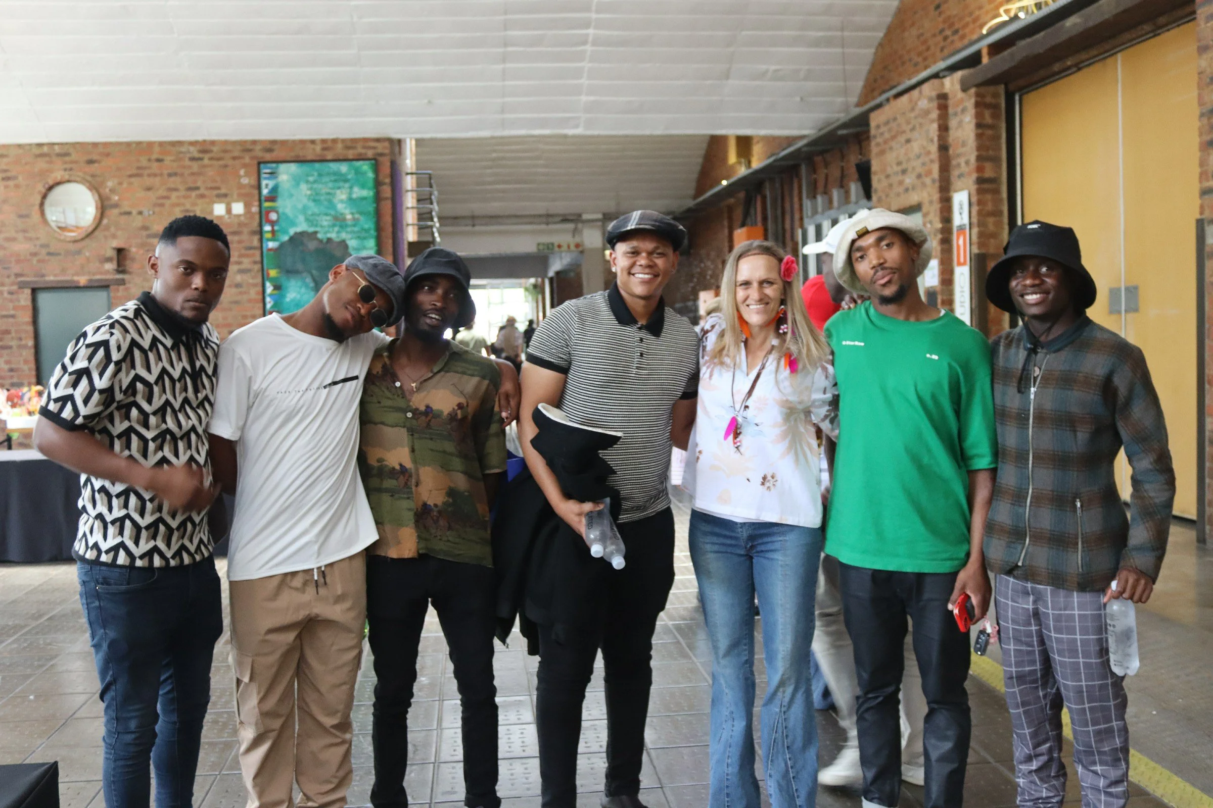 Group of seven diverse individuals standing together indoors, smiling, with brick walls and artwork in the background.