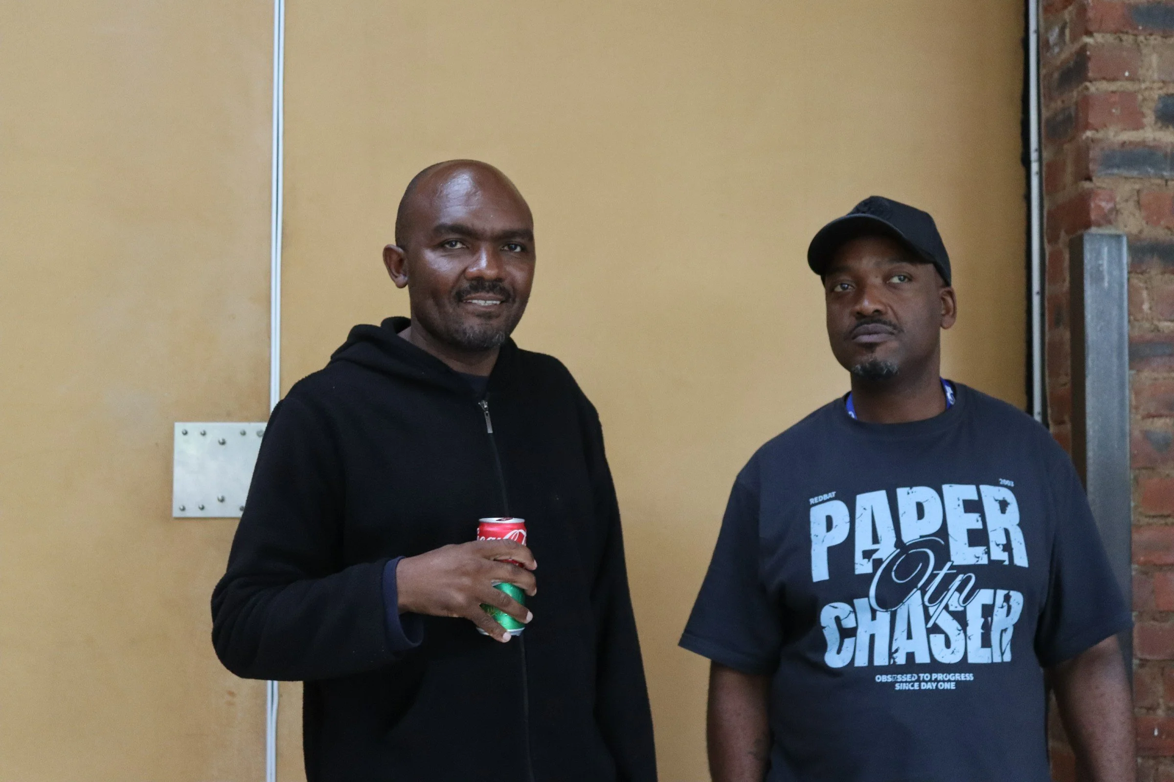 Two men standing indoors, one holding a can of soda, with a beige wall and exposed brickwork behind them.