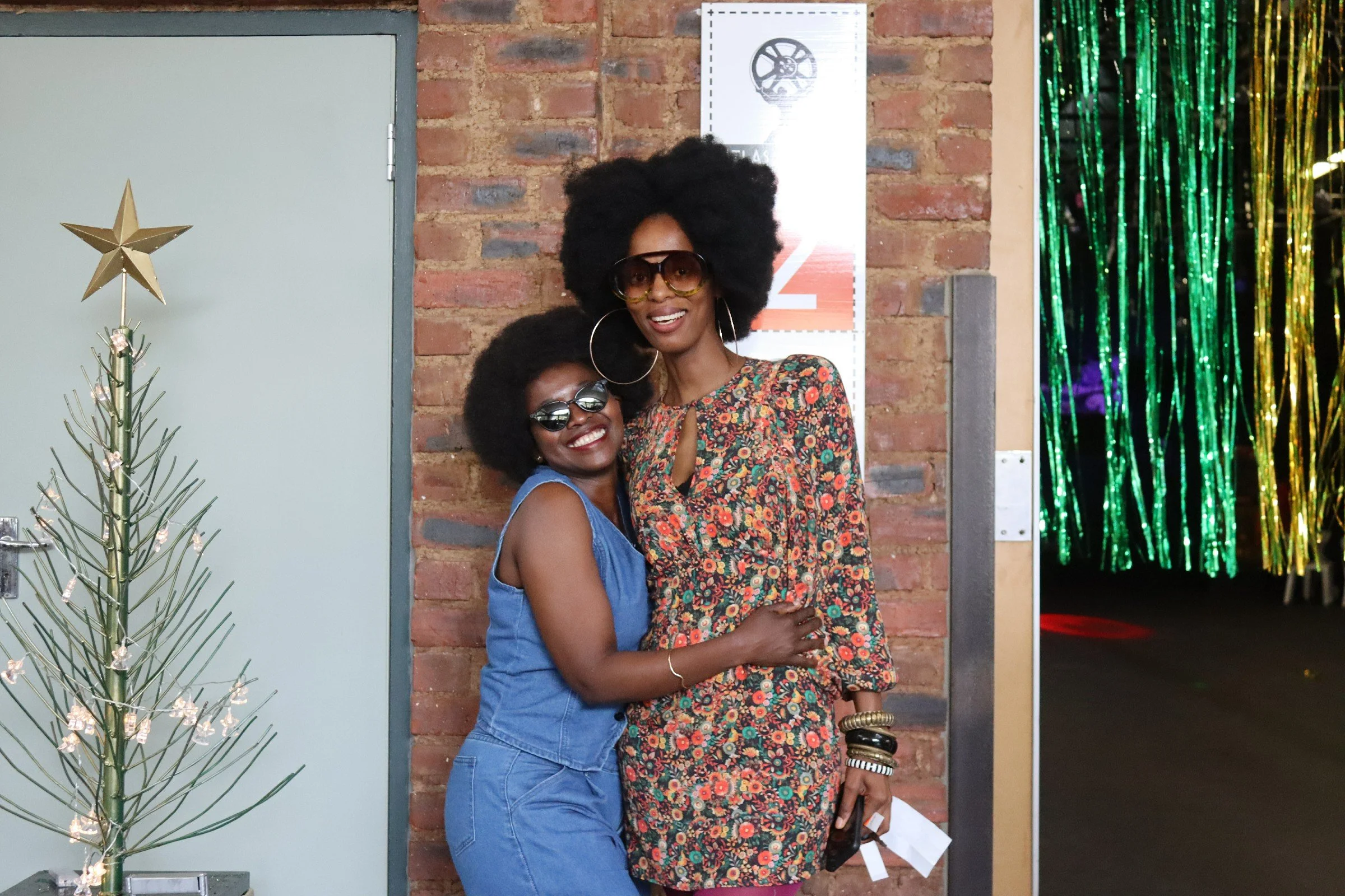 Two women with curly hair and glasses posing together, smiling in front of a brick wall decorated with a small Christmas tree and shiny green and gold streamers.