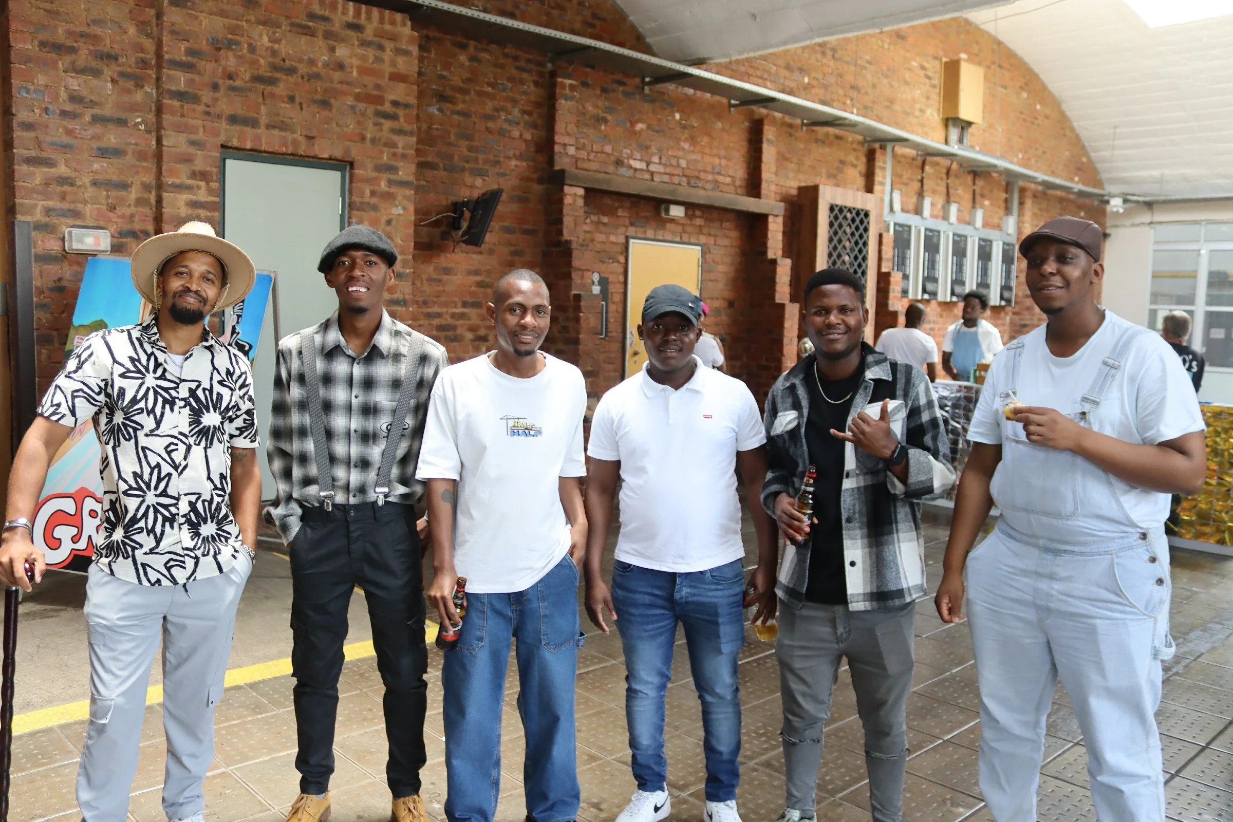 A group of seven men standing indoors in front of a brick wall and industrial shelves, smiling at the camera. Some are holding drinks, and they are dressed in casual attire including hats and t-shirts.