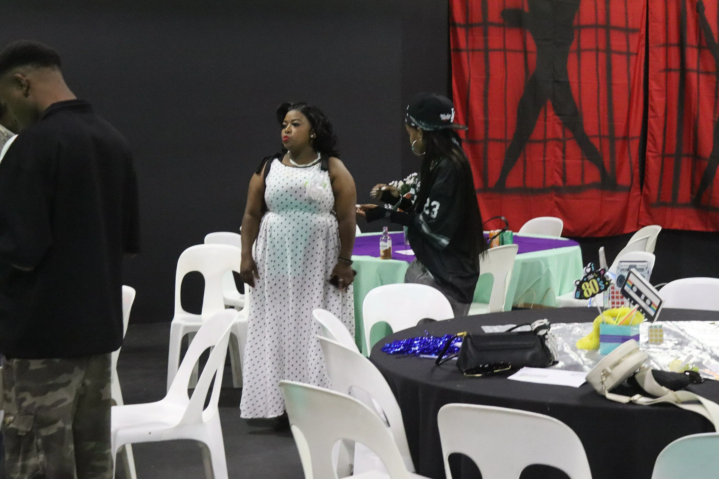 Three women standing at a decorated table in a celebration setting, with a red and black backdrop featuring a silhouette. One woman wears a polka dot dress, another a sporty outfit with a cap, and the third appears to be handling items on the table. 