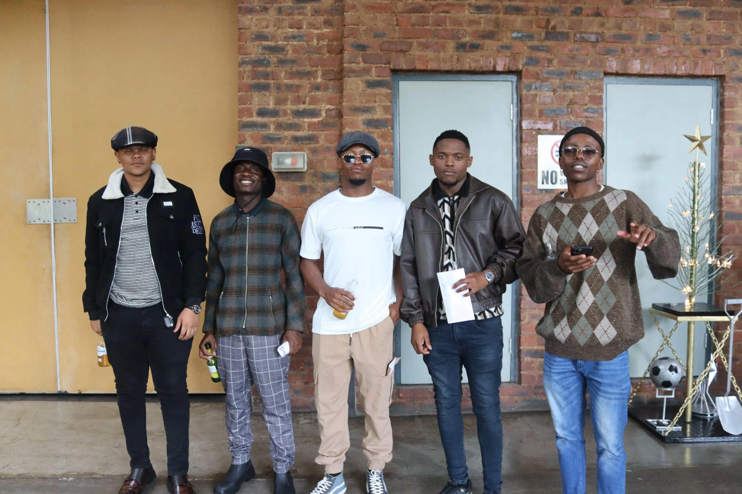 Five young men standing side by side indoors against a brick wall, some holding drinks, wearing casual clothing and accessories.