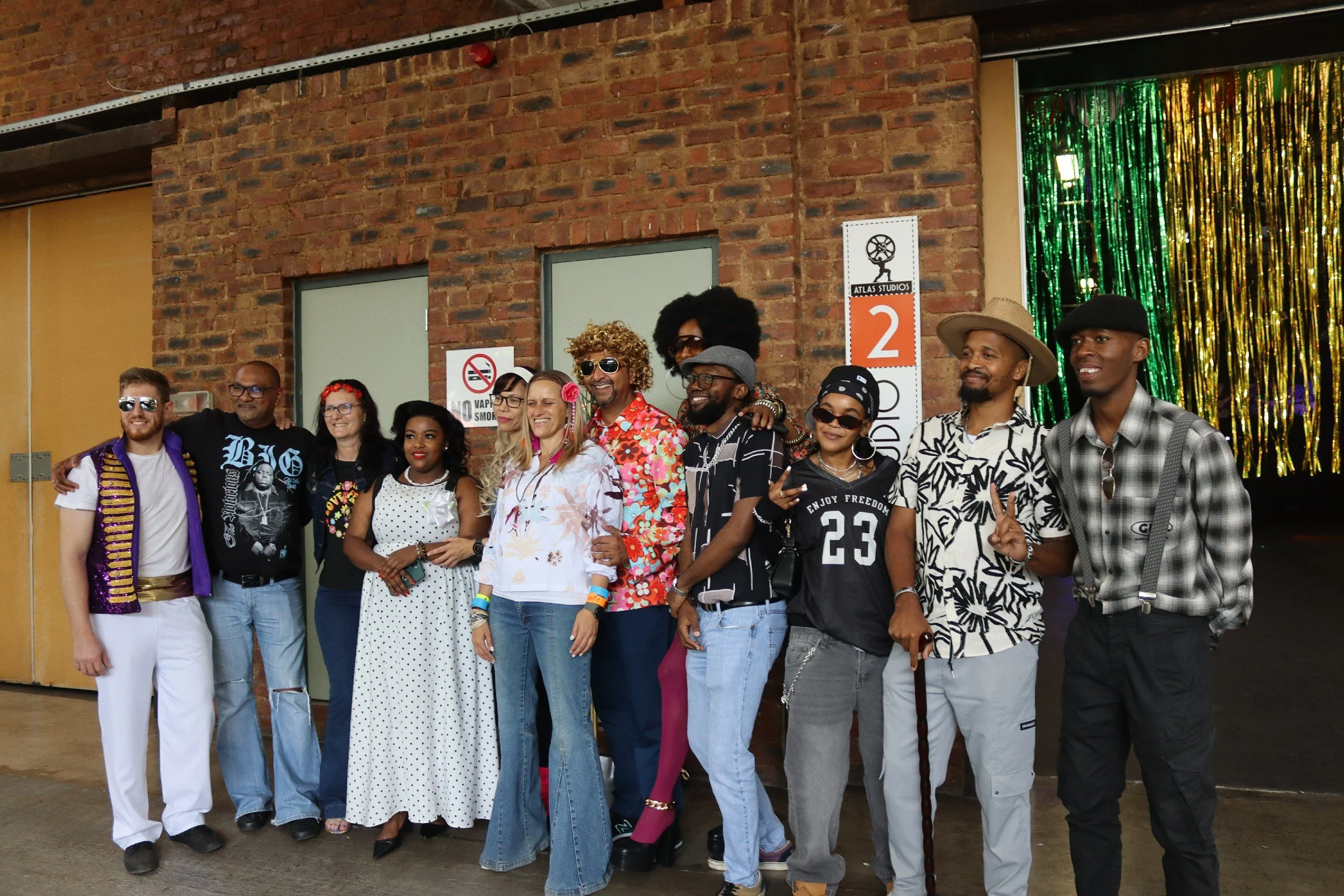 A diverse group of people standing together in front of an exposed brick wall, smiling and posing for a photo at an indoor event, with a sign reading 'Atlas Studios' and a colorful, shiny backdrop.