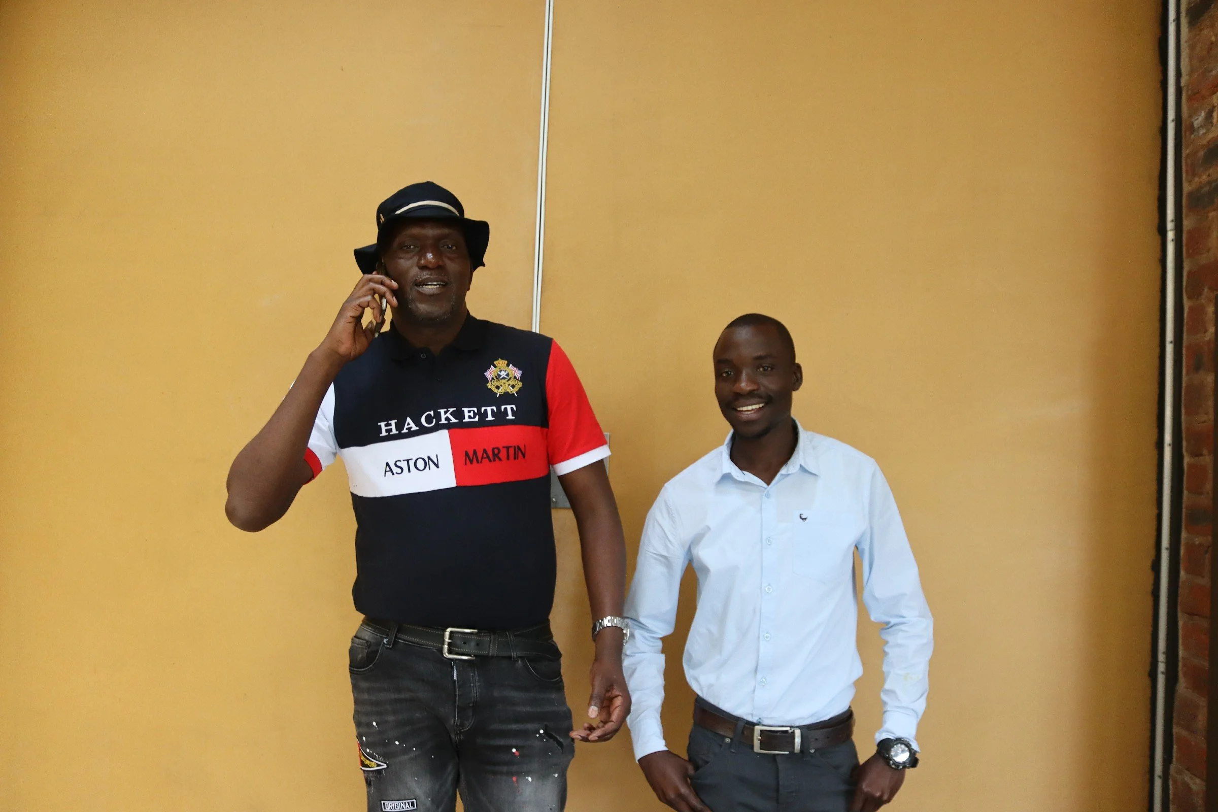 Two men standing indoors against a yellow wall, one talking on a mobile phone and the other smiling, holding hands.