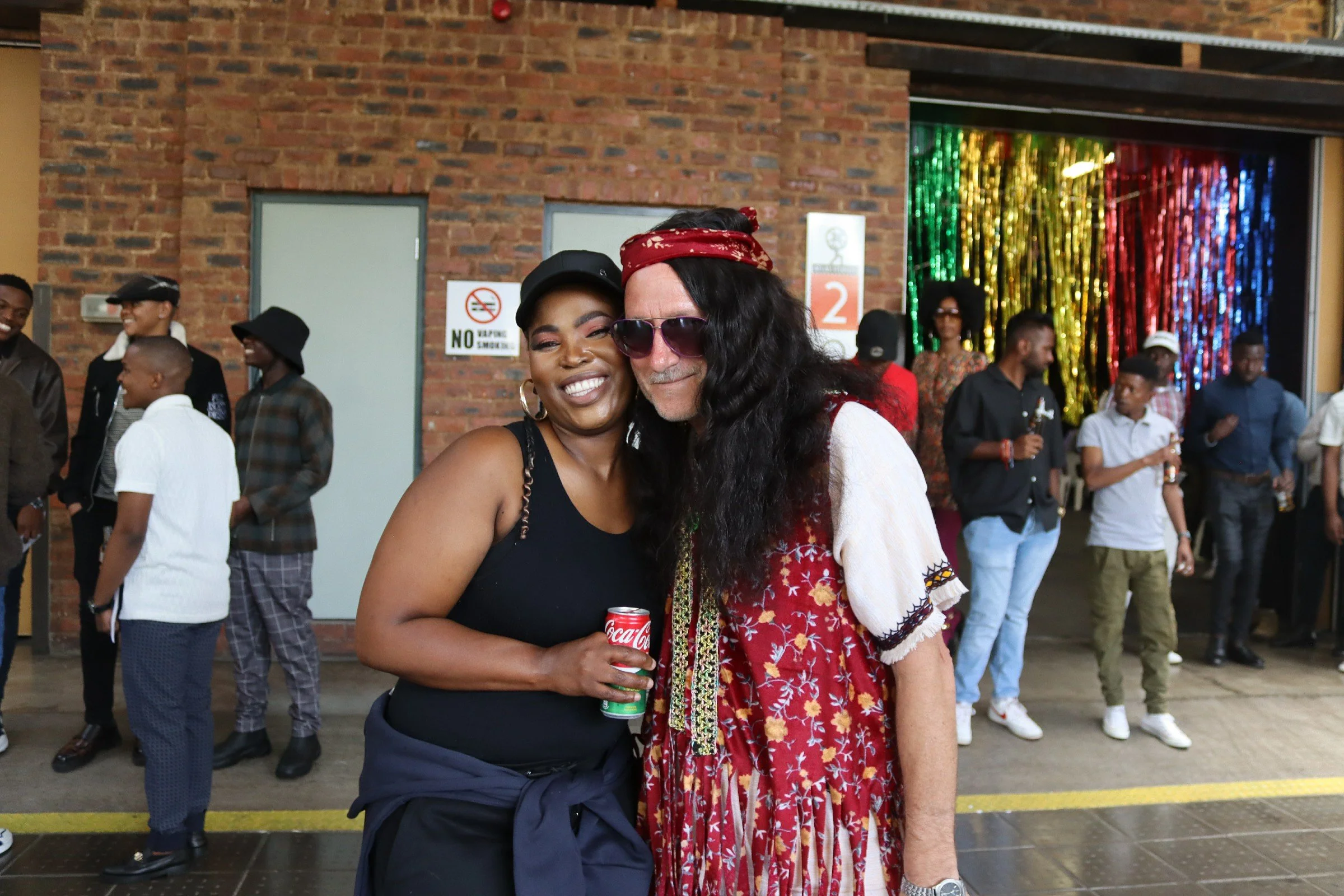 Two women hugging and smiling at an indoor event, with people in the background, brick walls, and colorful decorations.