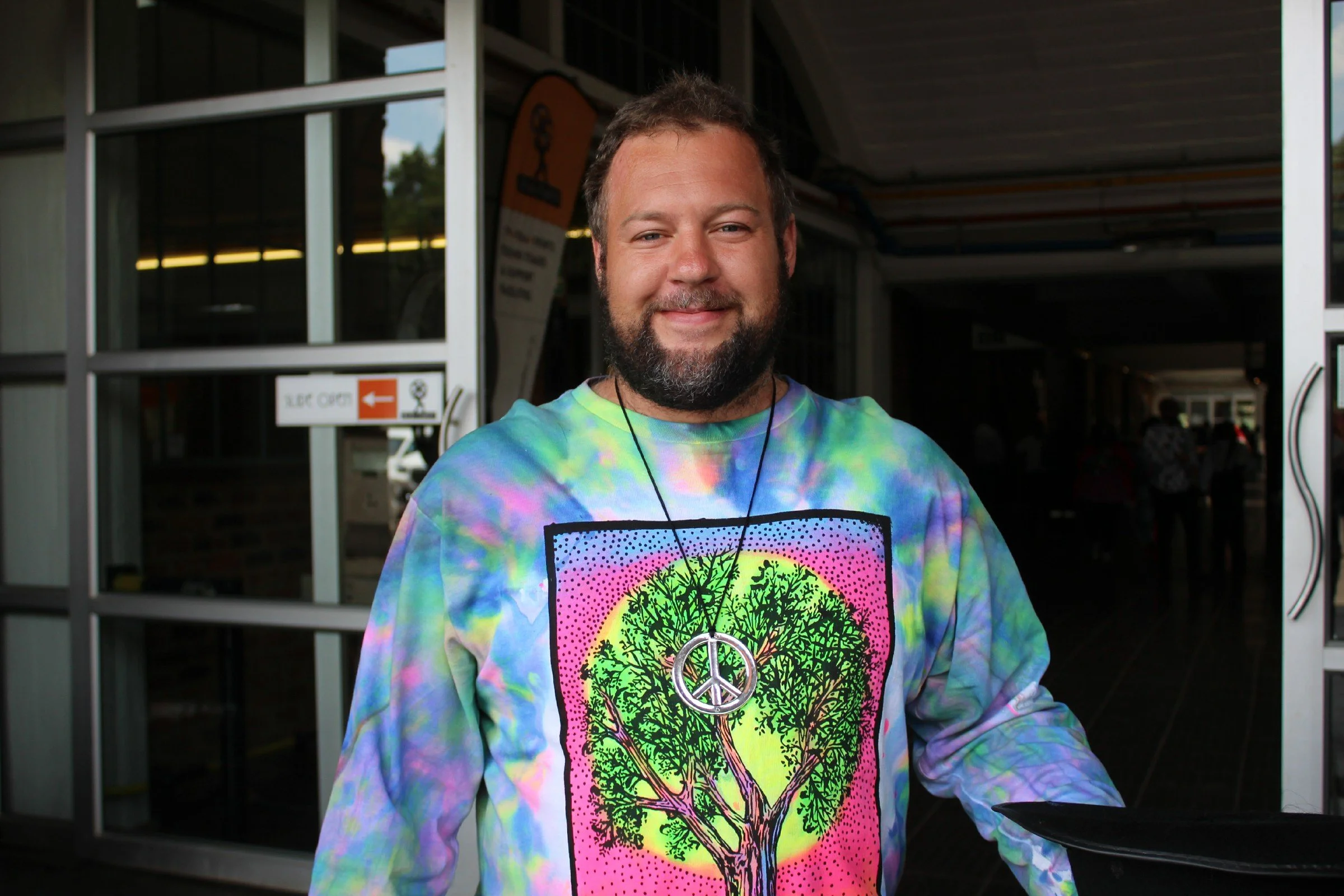 A man with a beard and short hair smiling, wearing a colorful tie-dye shirt with a peace symbol necklace and a peace sign on his shirt, standing in front of a building entrance with glass doors and a sign in the background.