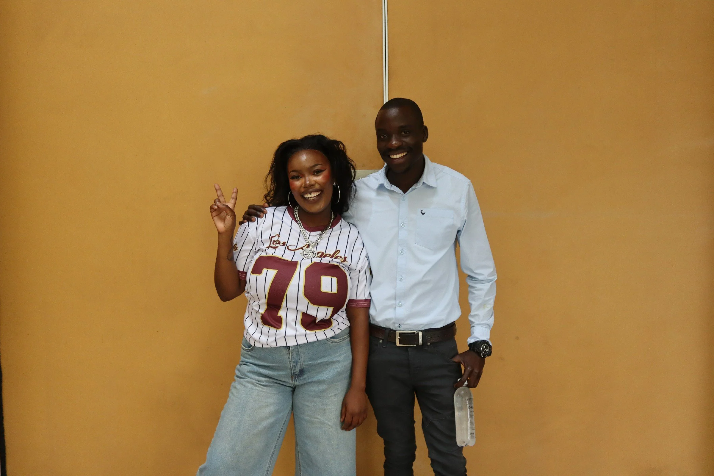 Two smiling individuals, a woman on the left with curly hair wearing a sports jersey and a man on the right with a shaved head wearing a white dress shirt, stand side by side against a beige wall, posing with the woman making a peace sign.