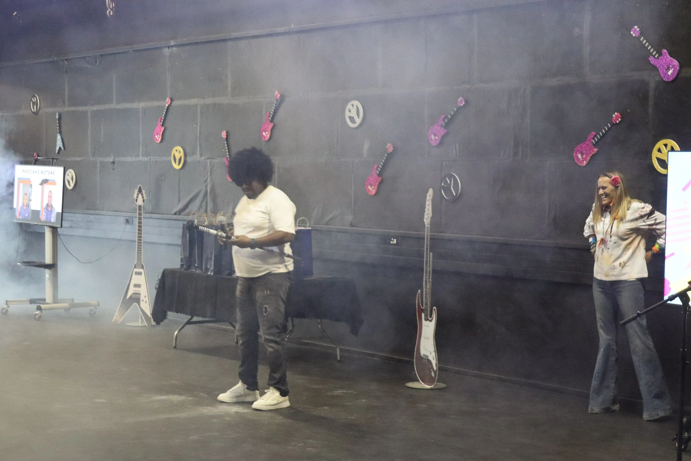 Indoor stage with two individuals, one man wearing sunglasses and casual clothes standing near musical instruments including electric guitars, and a woman with a flower in her hair standing nearby smiling. Decor on the wall includes pink glittery gui
