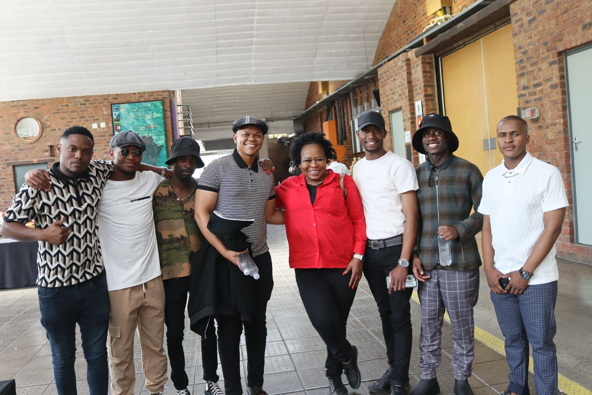 A group of eight people standing together indoors, smiling, with a brick wall and a yellow door in the background.