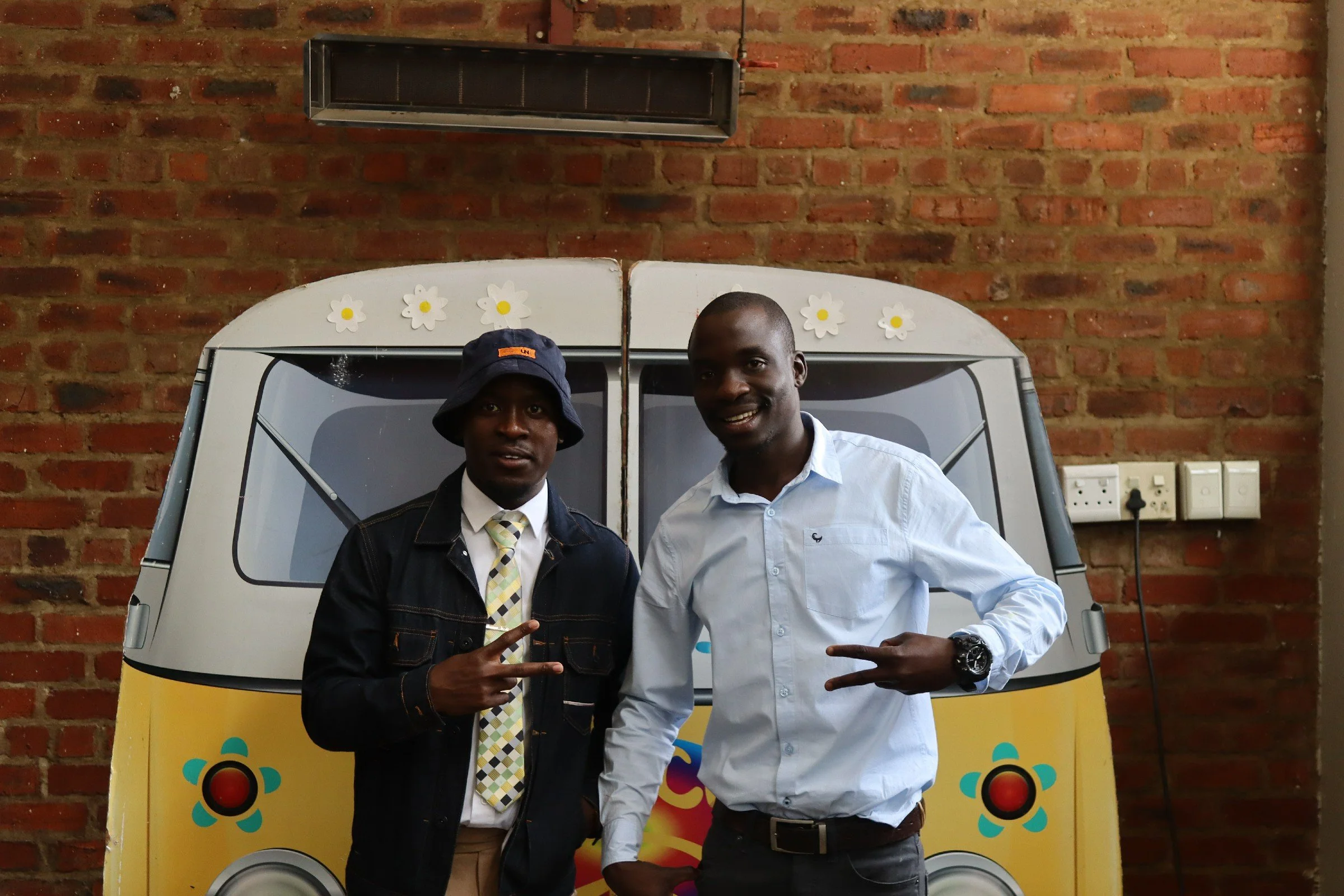 Two men standing in front of a graffiti-covered van with a brick wall background. One man is wearing a dark jacket, a patterned tie, and a bucket hat, and is making a peace sign with his hand. The other man is dressed in a light blue shirt and smilin