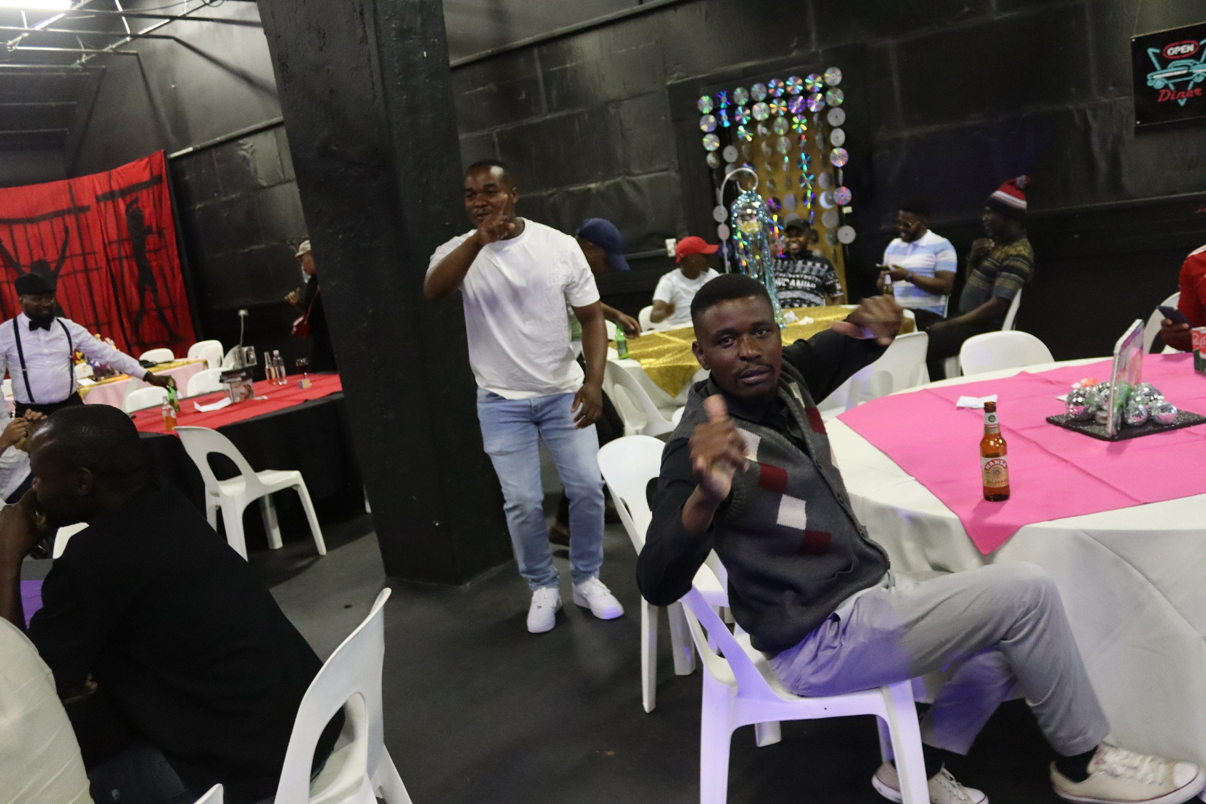 People gathered around tables with pink tablecloths in a party room, some sitting and some standing, with decorations including a dress form ornament and a red wall hanging in the background. One man in a black jacket is giving a thumbs-up while seat