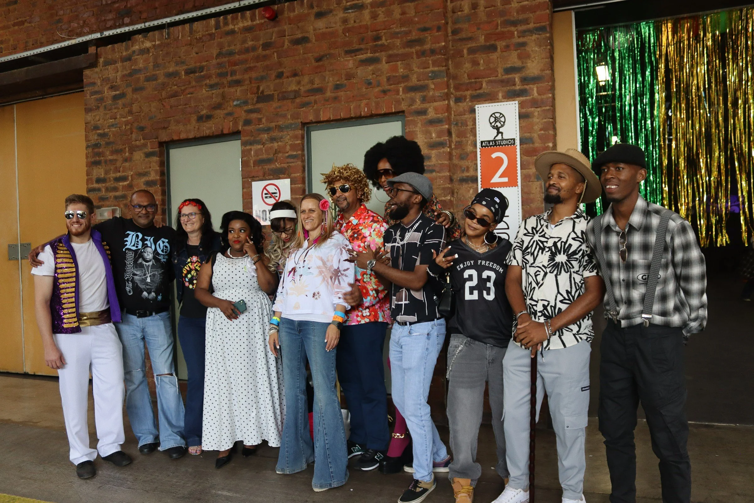 A group of diverse people dressed in colorful and themed outfits, standing together in front of a brick wall with signs, some wearing sunglasses, smiling and posing for a photo at an event.